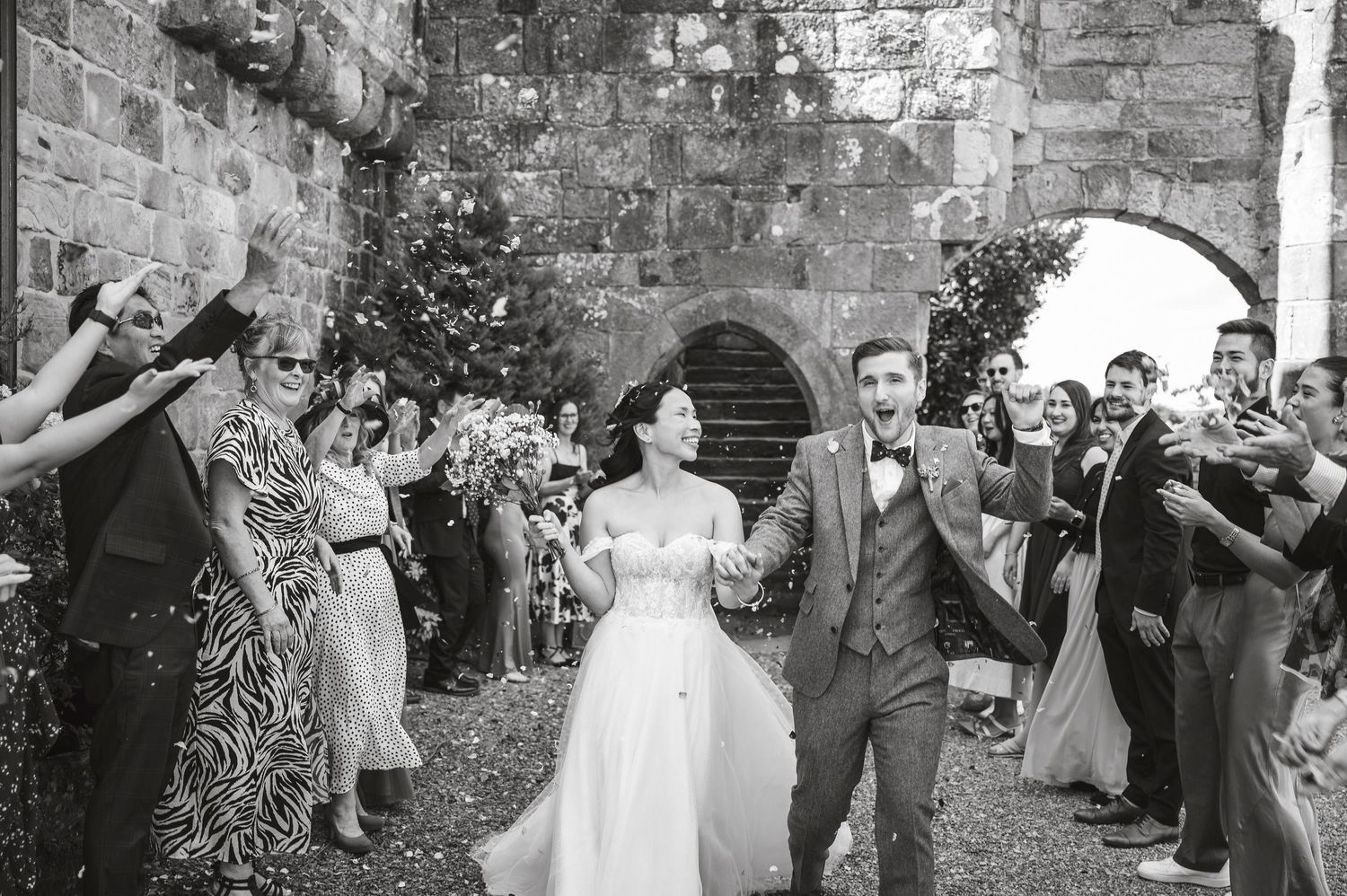 Wedding celebration with guests throwing confetti in front of stone archway.