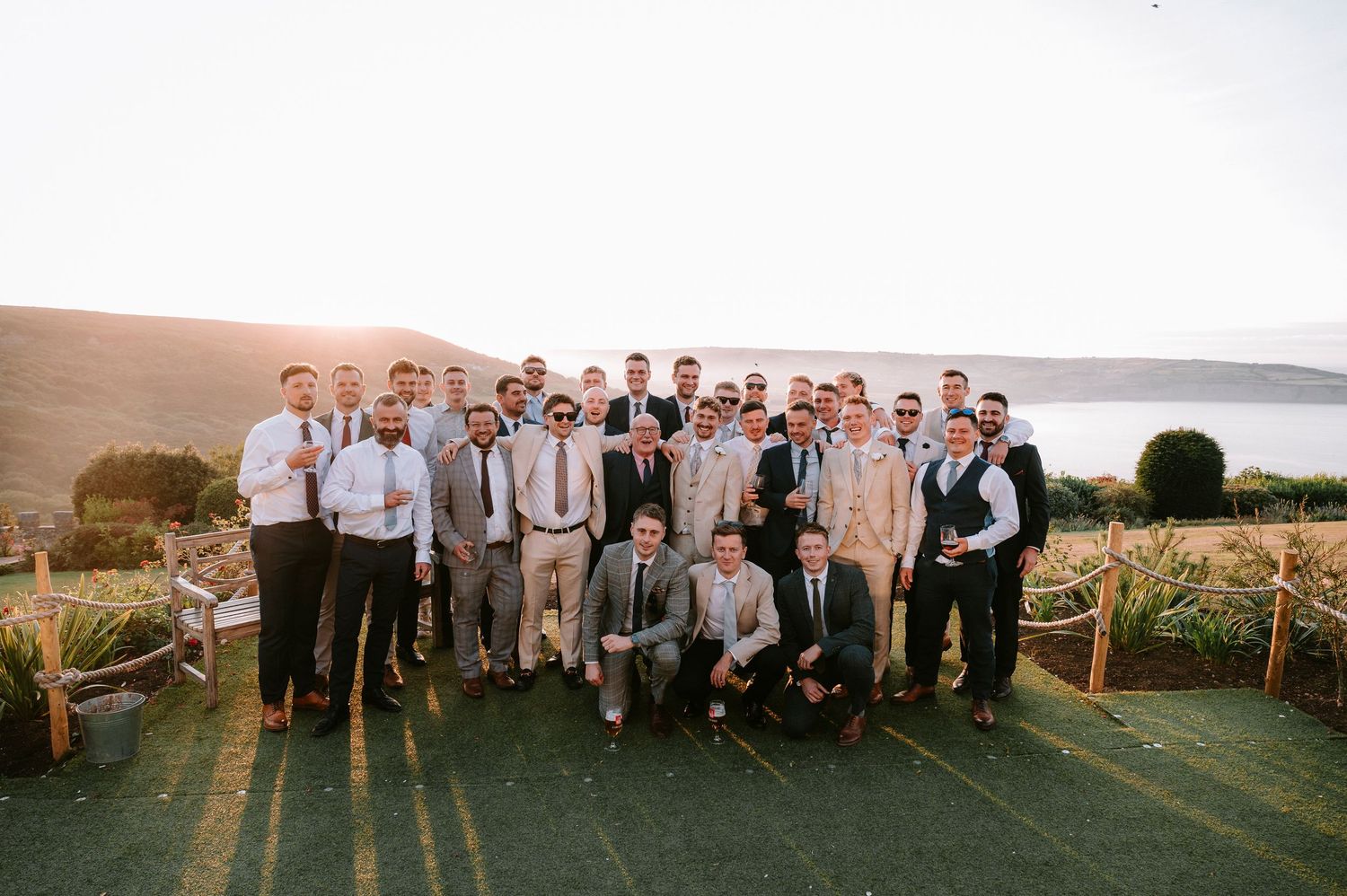 Large wedding party group photo with groomsmen and guests posed outdoors at sunset.