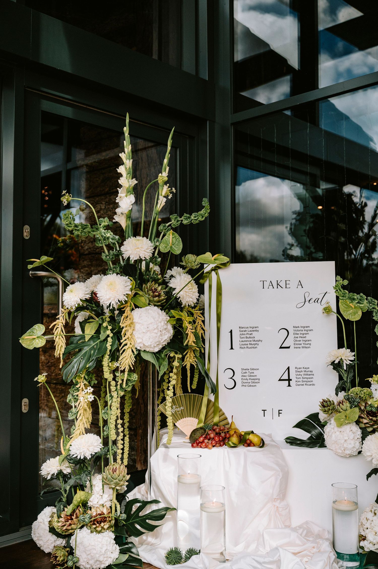 Wedding reception table displays with white floral arrangements, candles, and seating chart numbers against dark windows.