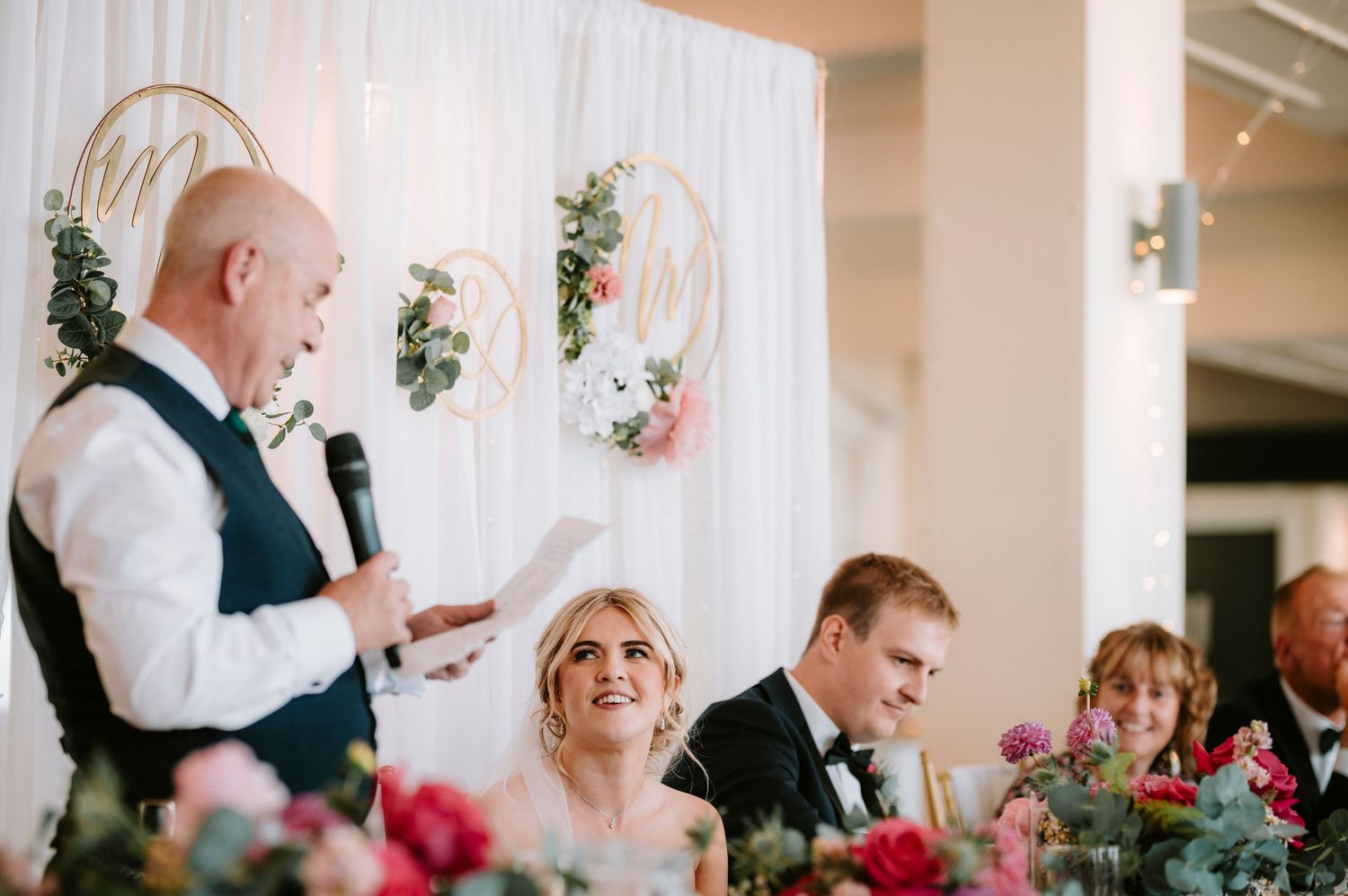 Wedding reception speeches at decorated head table with floral arrangements.