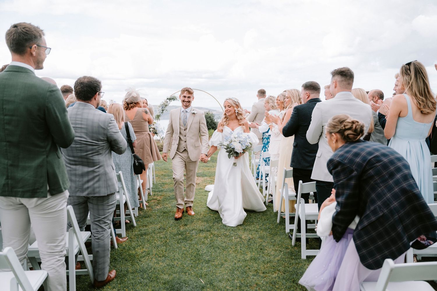 Wedding ceremony guests stand and celebrate on a grassy lawn with white chairs.