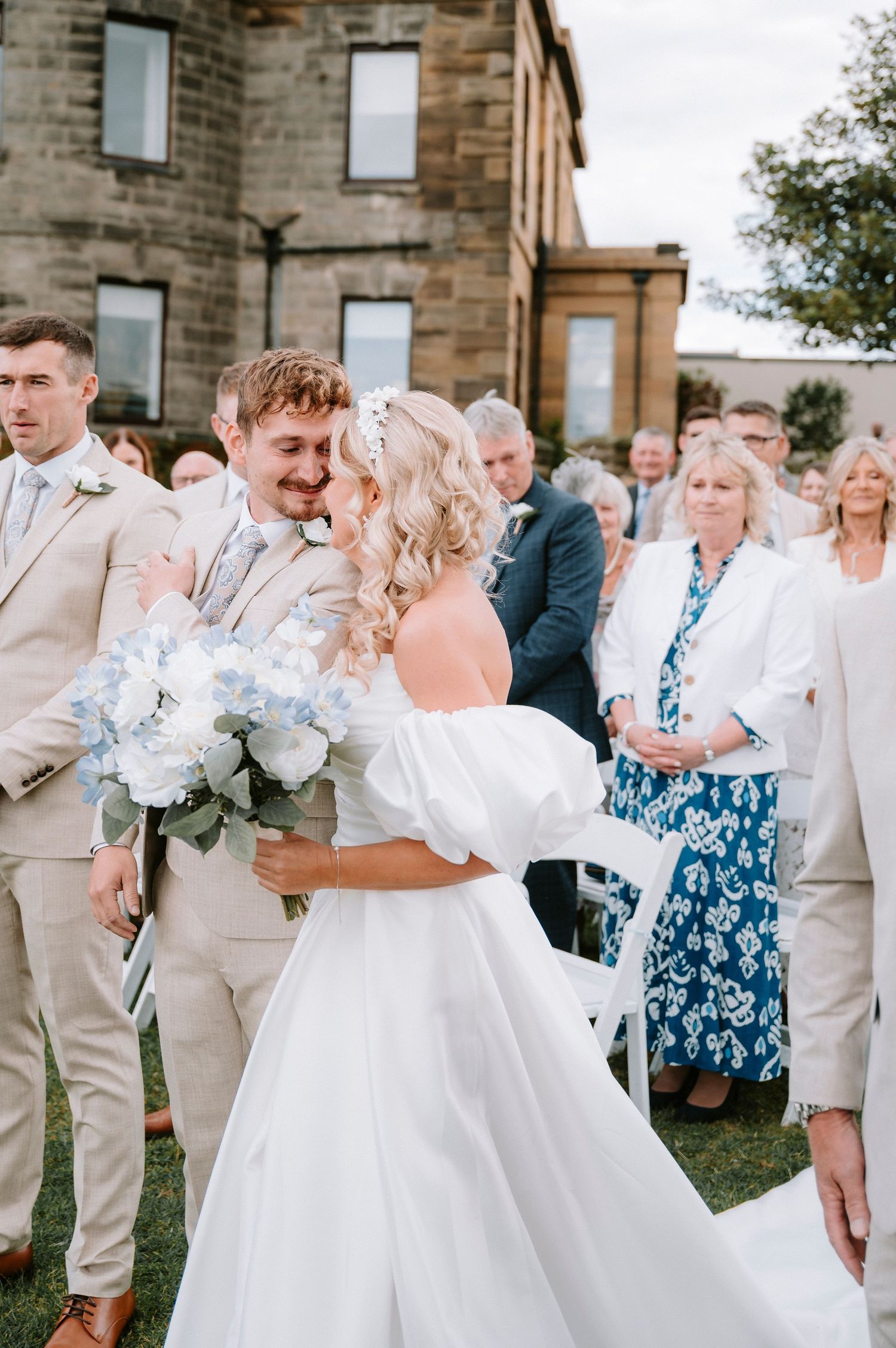 Wedding party celebrating outdoors with guests in beige suits and blue floral dresses.