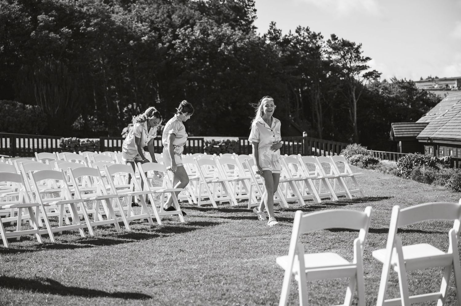 Black and white photo of wedding party walking through rows of white chairs on lawn.