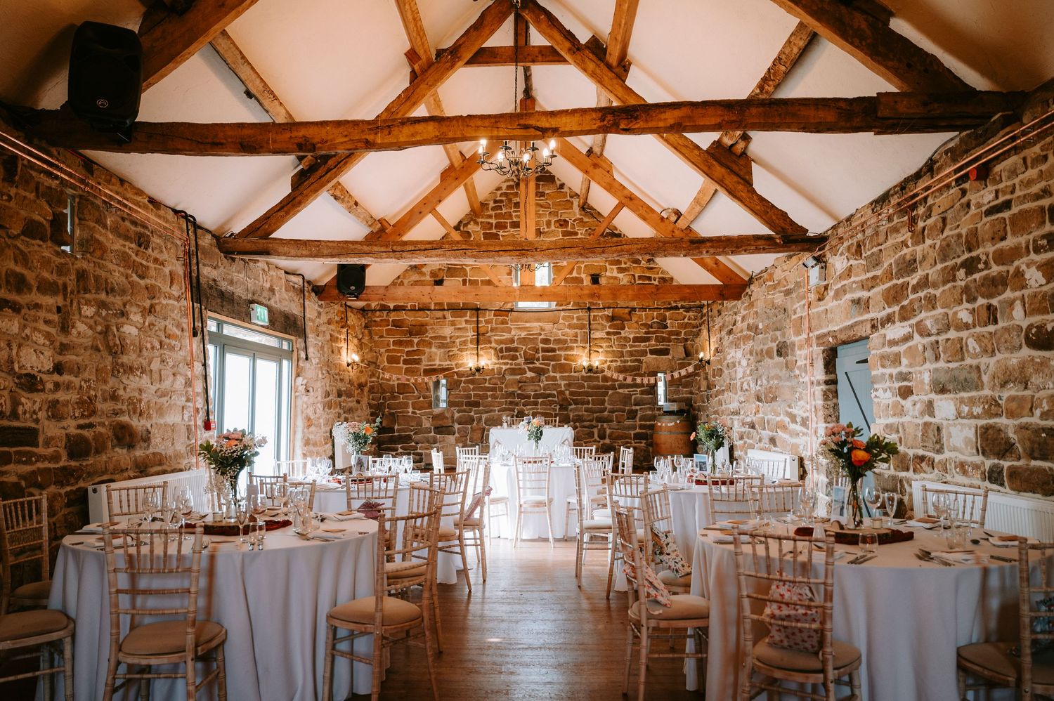 Rustic barn wedding reception venue with exposed wooden beams and stone walls set with round tables.