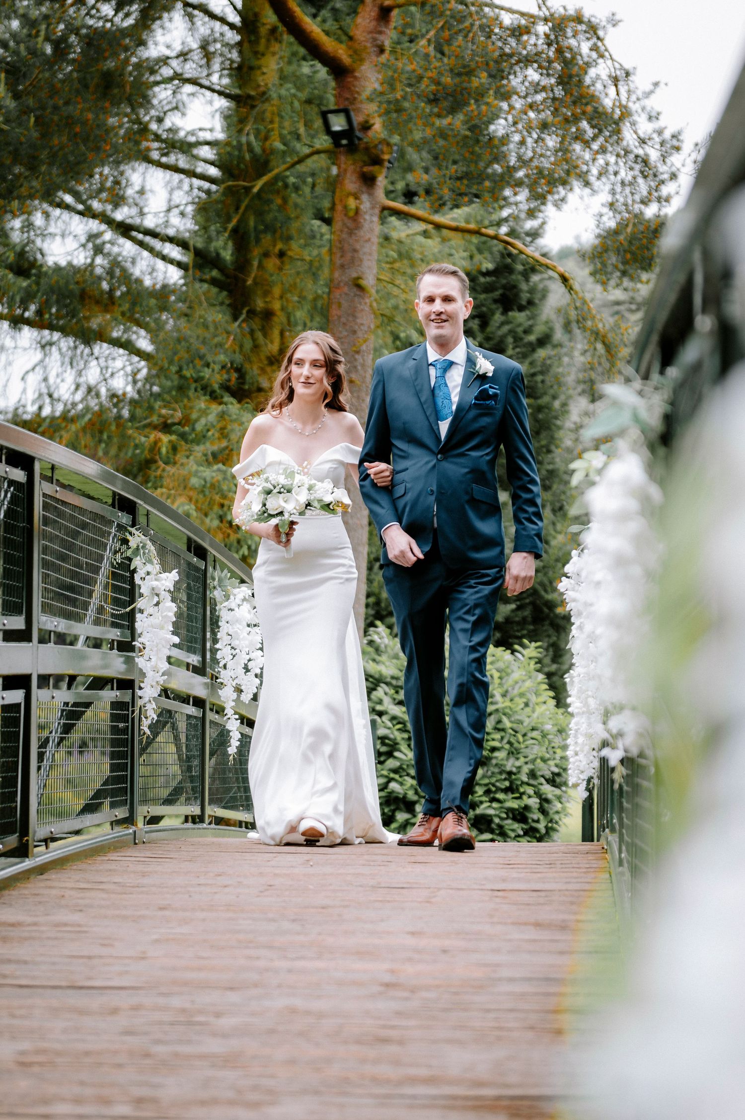 Wedding couple walking along a modern wooden bridge decorated with white floral arrangements.