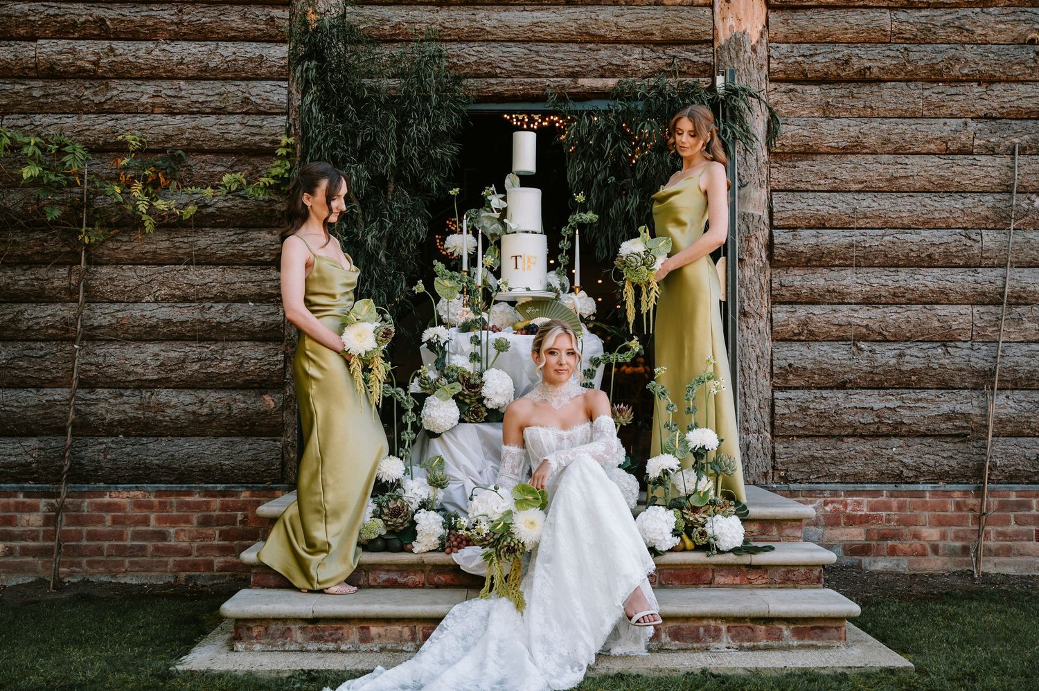 A wedding party poses in front of a rustic log wall with bridesmaids in sage green dresses and white floral bouquets.