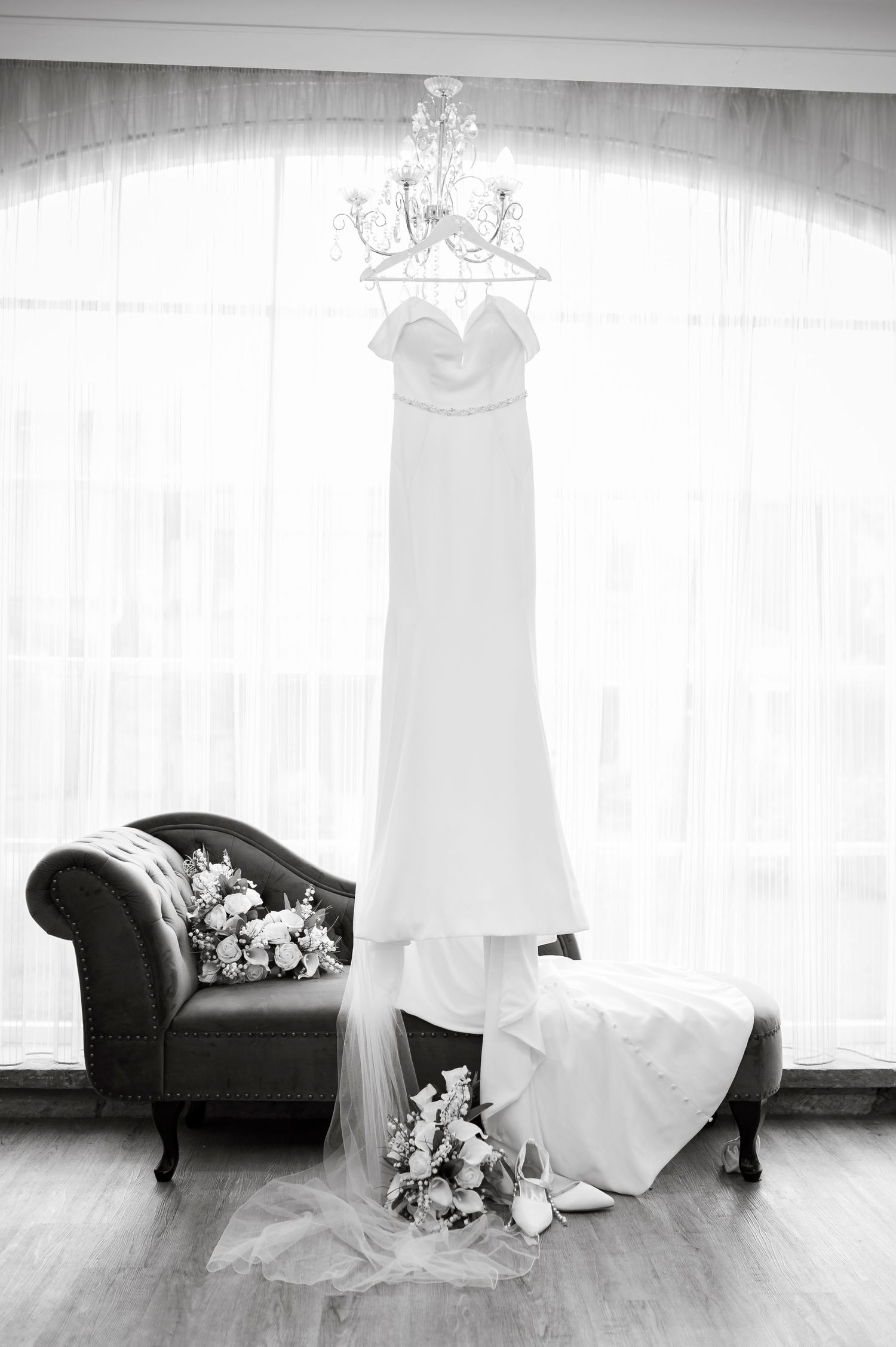 An elegant white wedding dress hangs from a chandelier above a vintage chaise lounge.