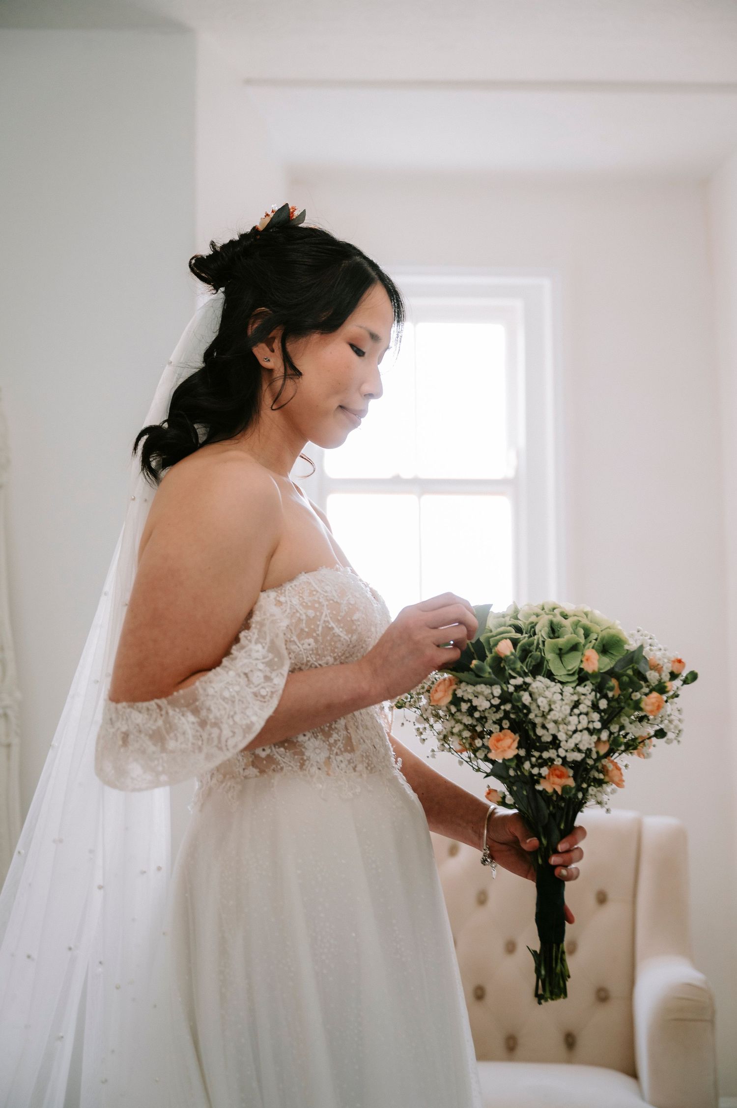 Bride in a lace wedding dress holding a peach and white floral bouquet in a bright white room.
