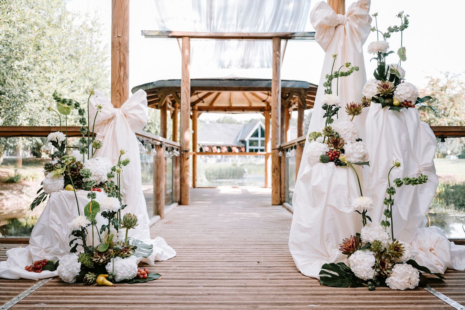 White fabric draped wedding arch decorated with roses and greenery on wooden deck pathway in outdoor garden setting.