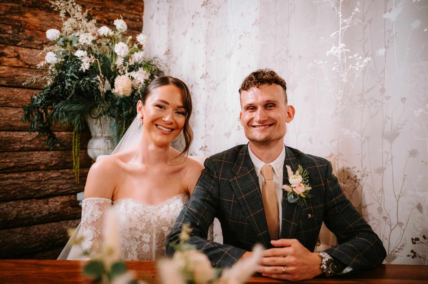 Newlyweds smile together at rustic wedding reception with floral arrangements and exposed brick walls.
