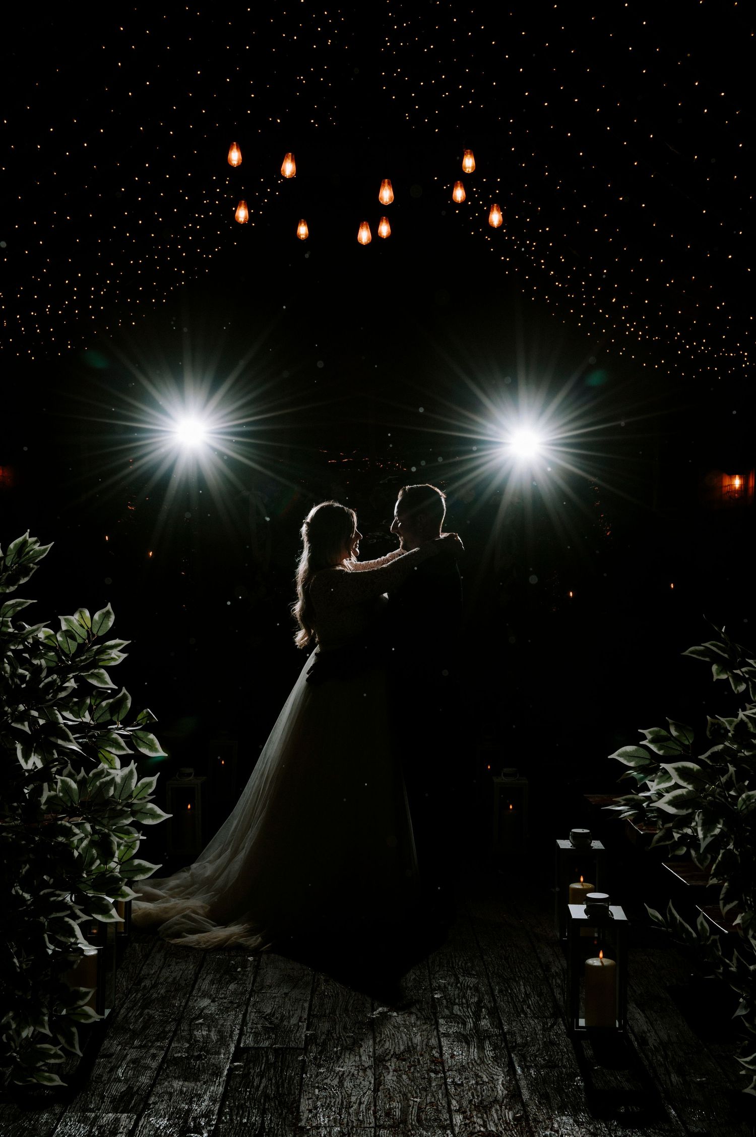 A dramatic nighttime wedding photo with silhouettes against bright spotlights and twinkling lights.