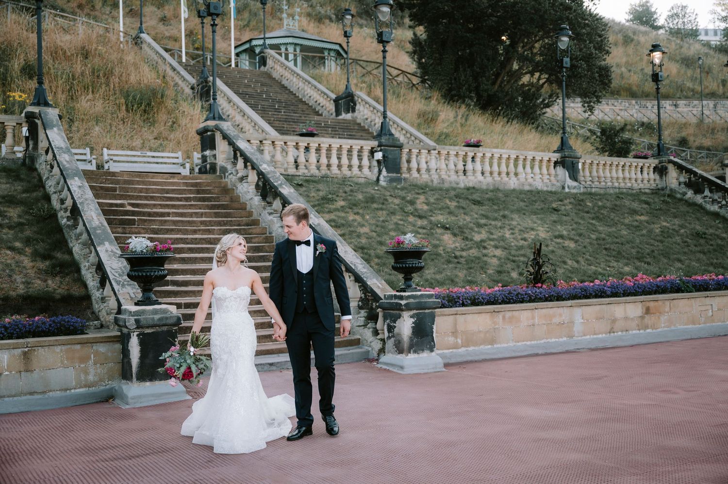 Couple walking along stone balustrade pathway during wedding photoshoot.