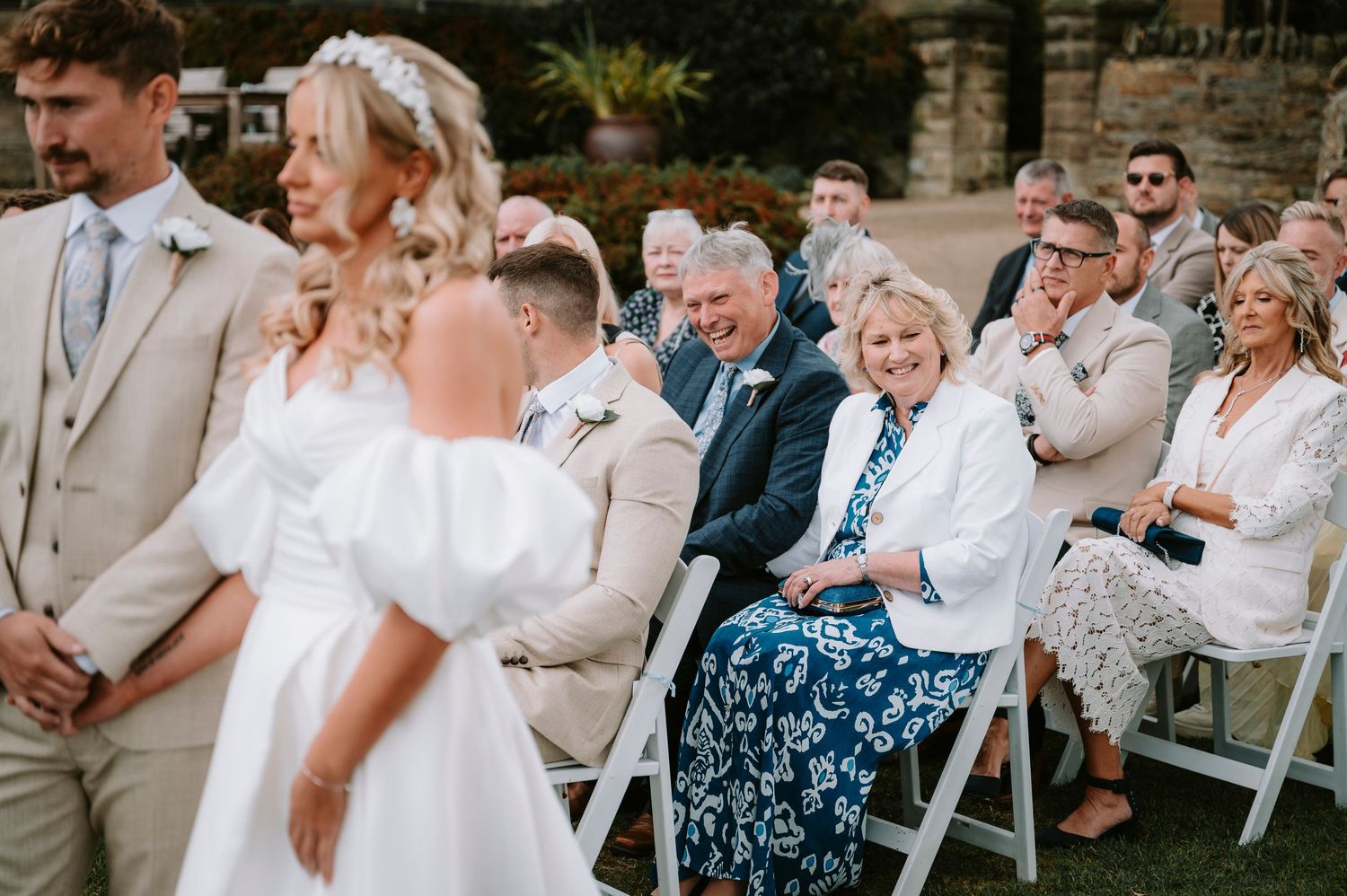 Wedding celebration outdoors with guests seated in white chairs near a stone building.