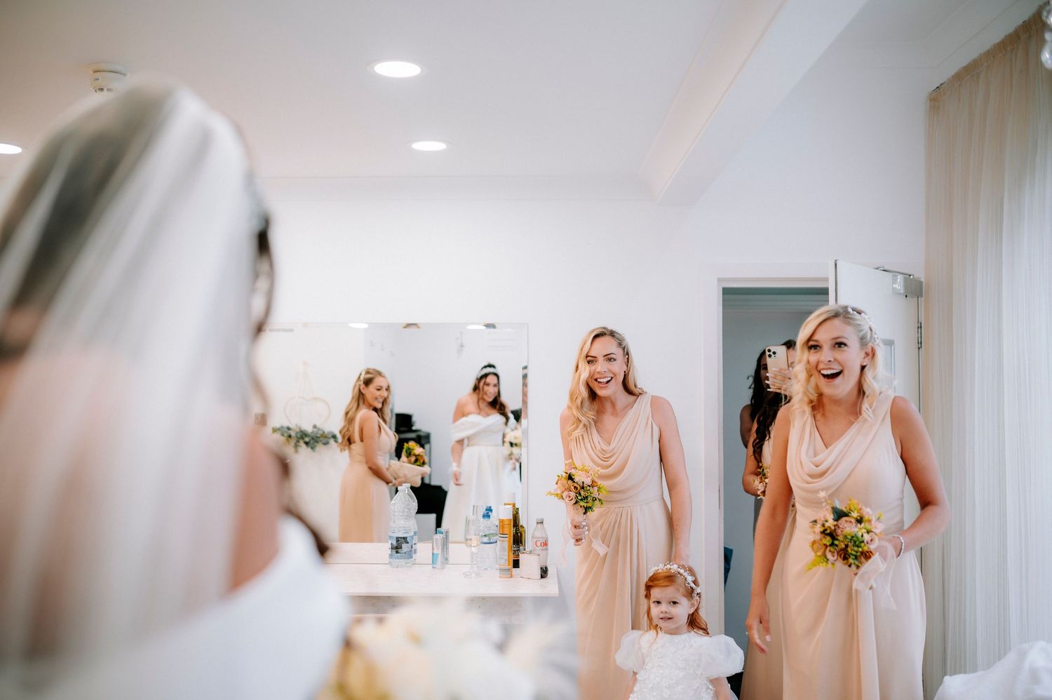Bridesmaids in blush pink dresses prepare for a wedding celebration in a bright indoor space.