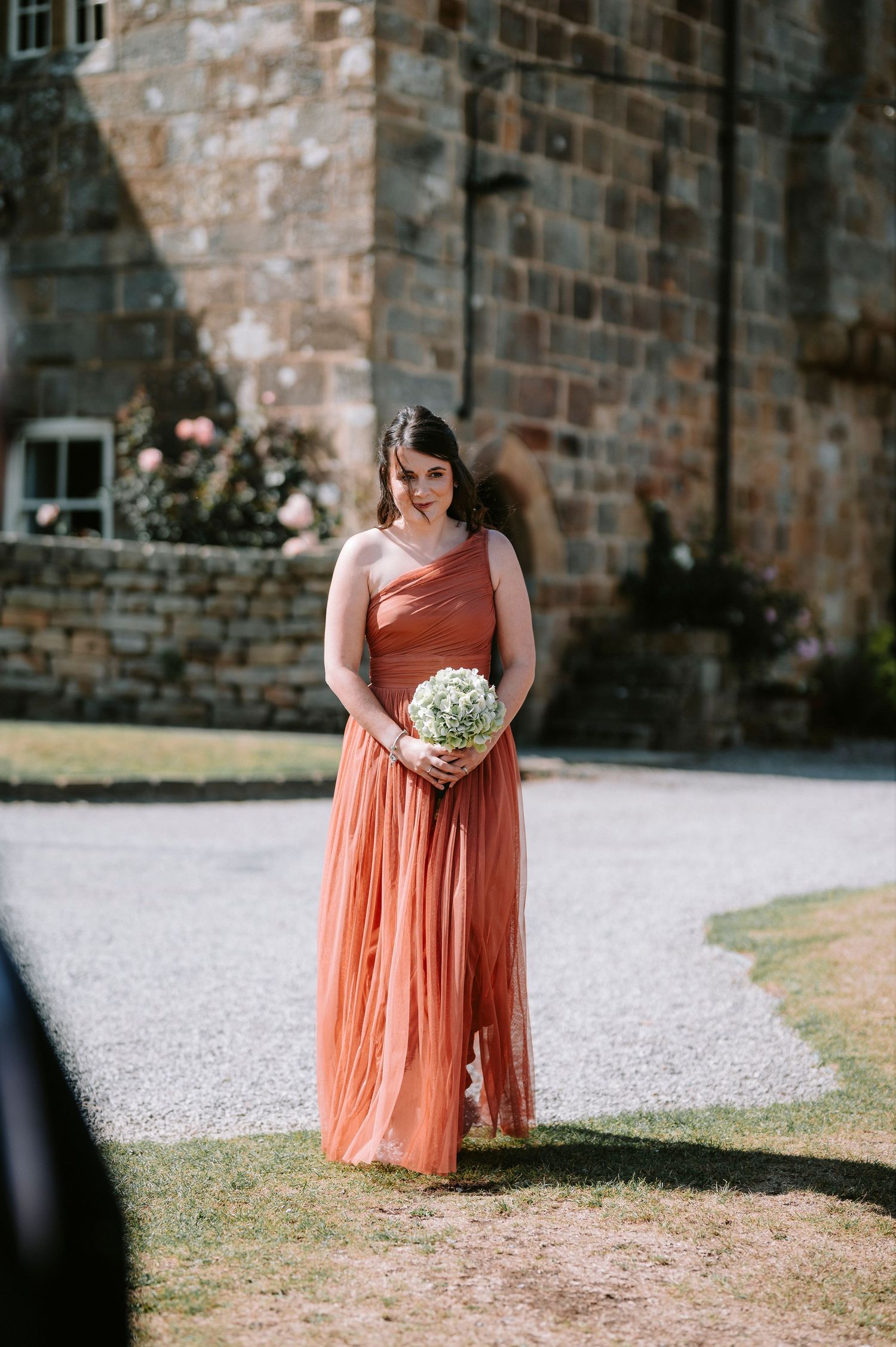 A bridesmaid in a flowing peach gown holds flowers against a rustic stone wall backdrop.