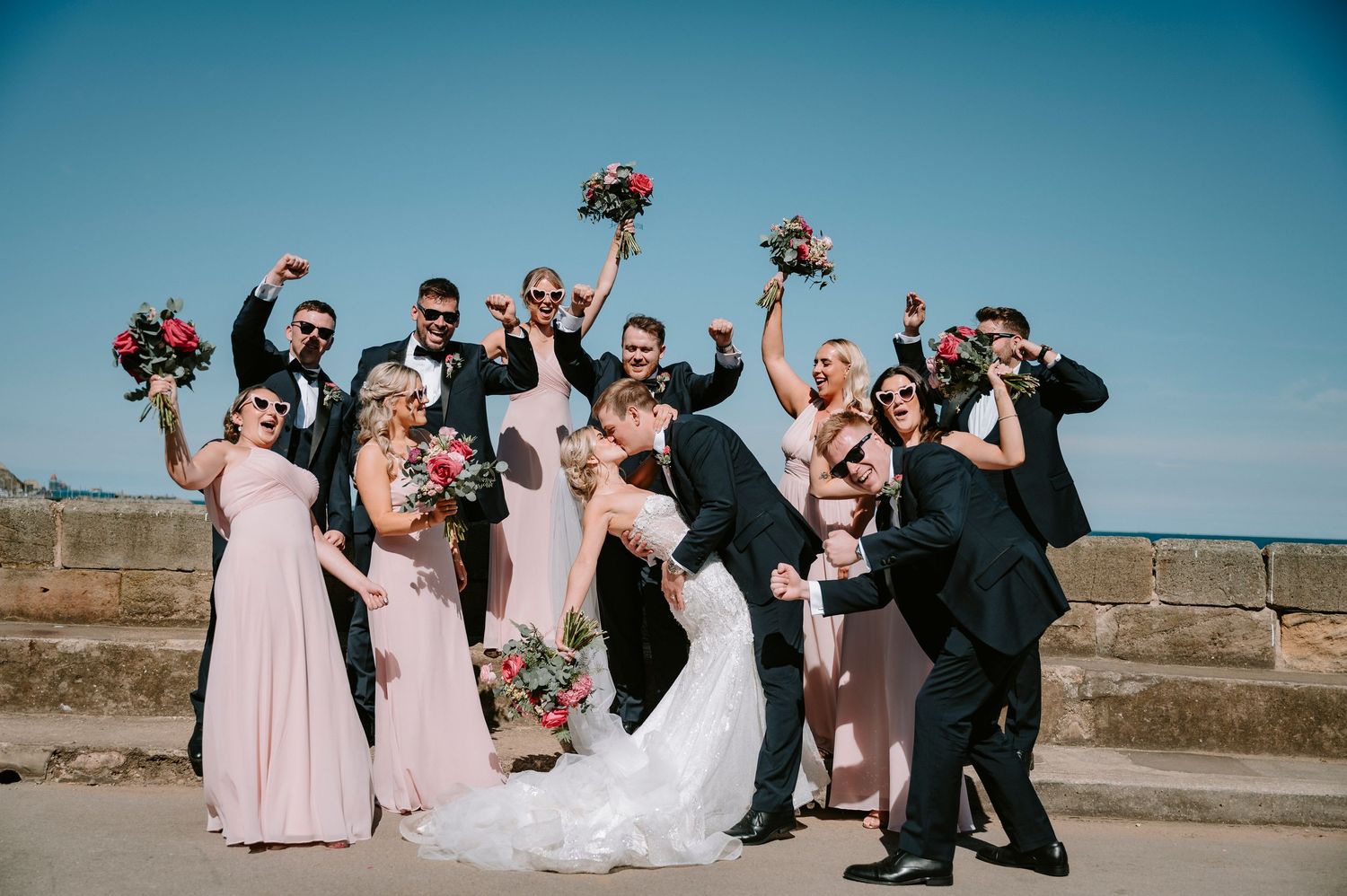 Wedding party celebrating joyfully outdoors with bridesmaids in pink and groomsmen in black suits.