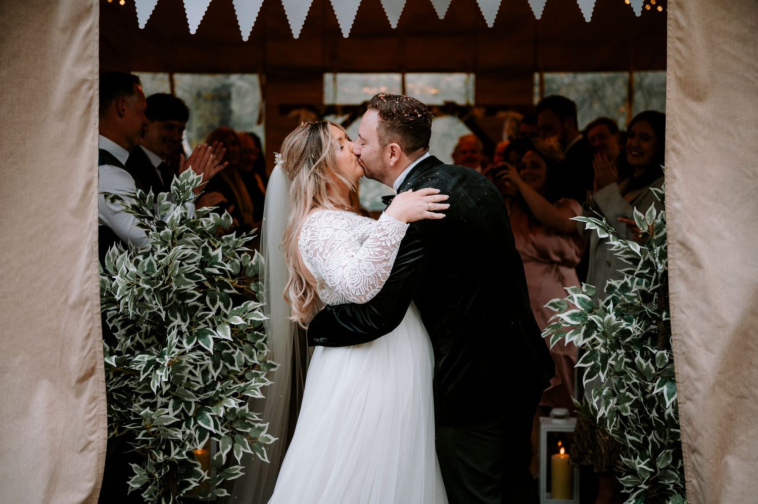 A romantic wedding kiss under decorative bunting surrounded by greenery and guests at an indoor venue.