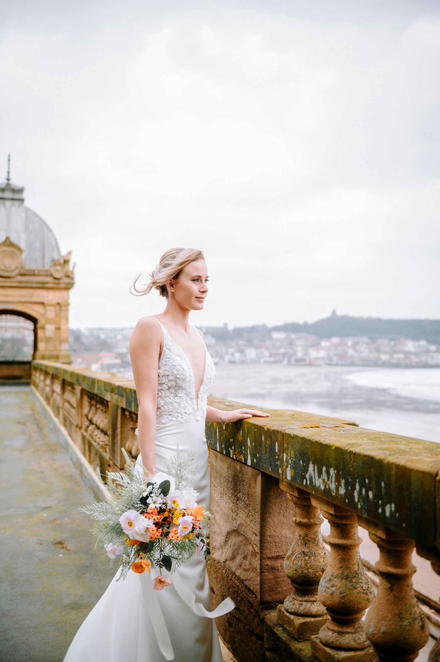 White lace wedding dress and bouquet against stone balustrade with ocean view backdrop.