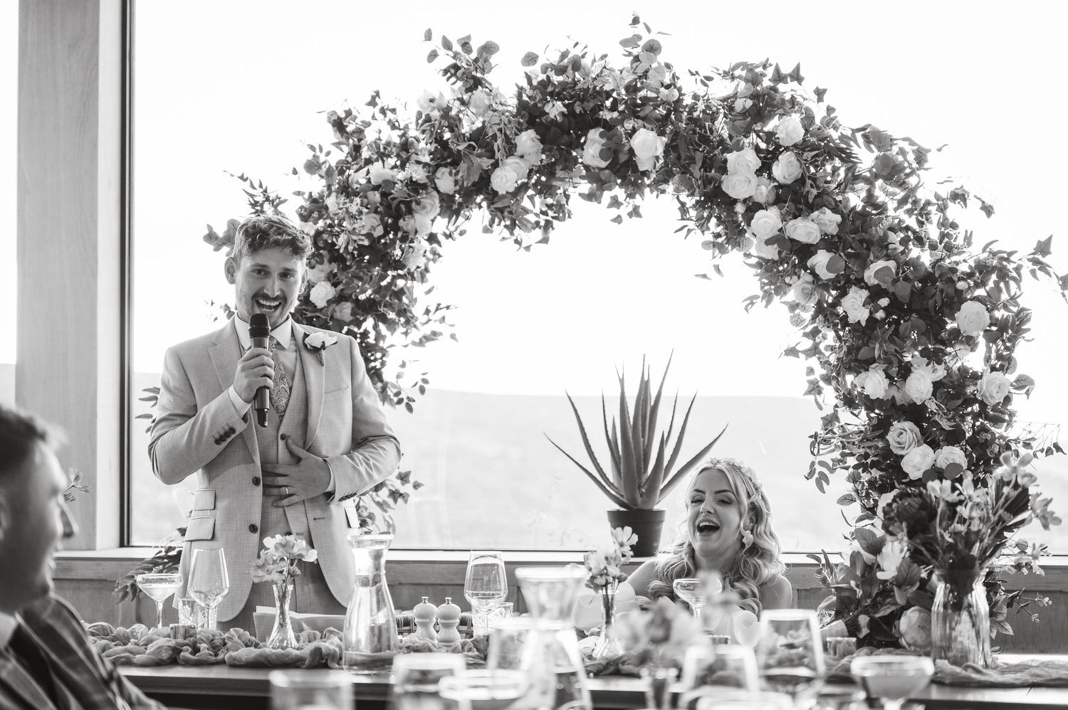 A wedding reception speech under an elegant floral arch with ocean views in the background.