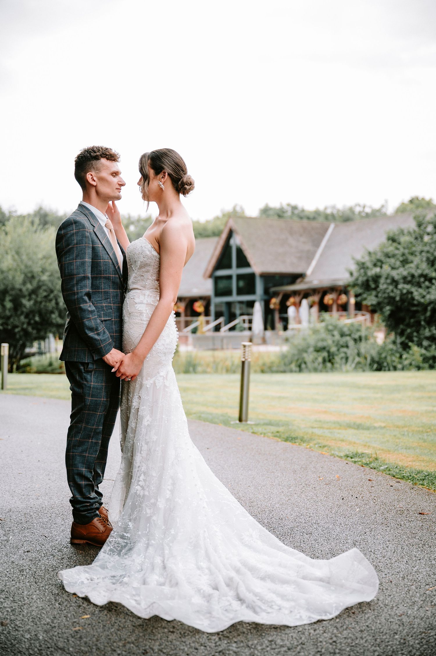 Bride in lace gown and groom in plaid suit pose outside rustic wedding venue.