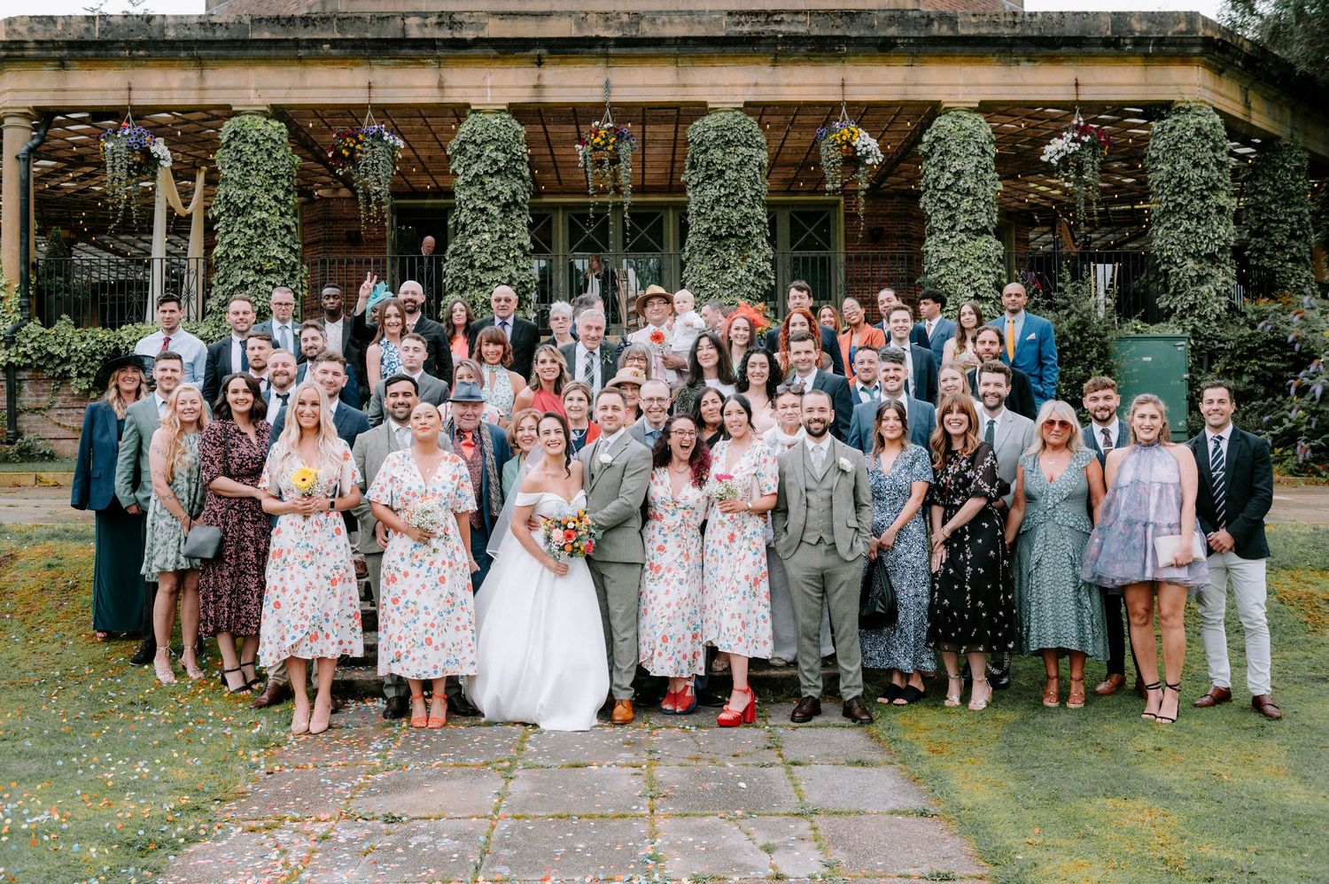 Large wedding party group photo with bridesmaids and groomsmen in outdoor garden setting.
