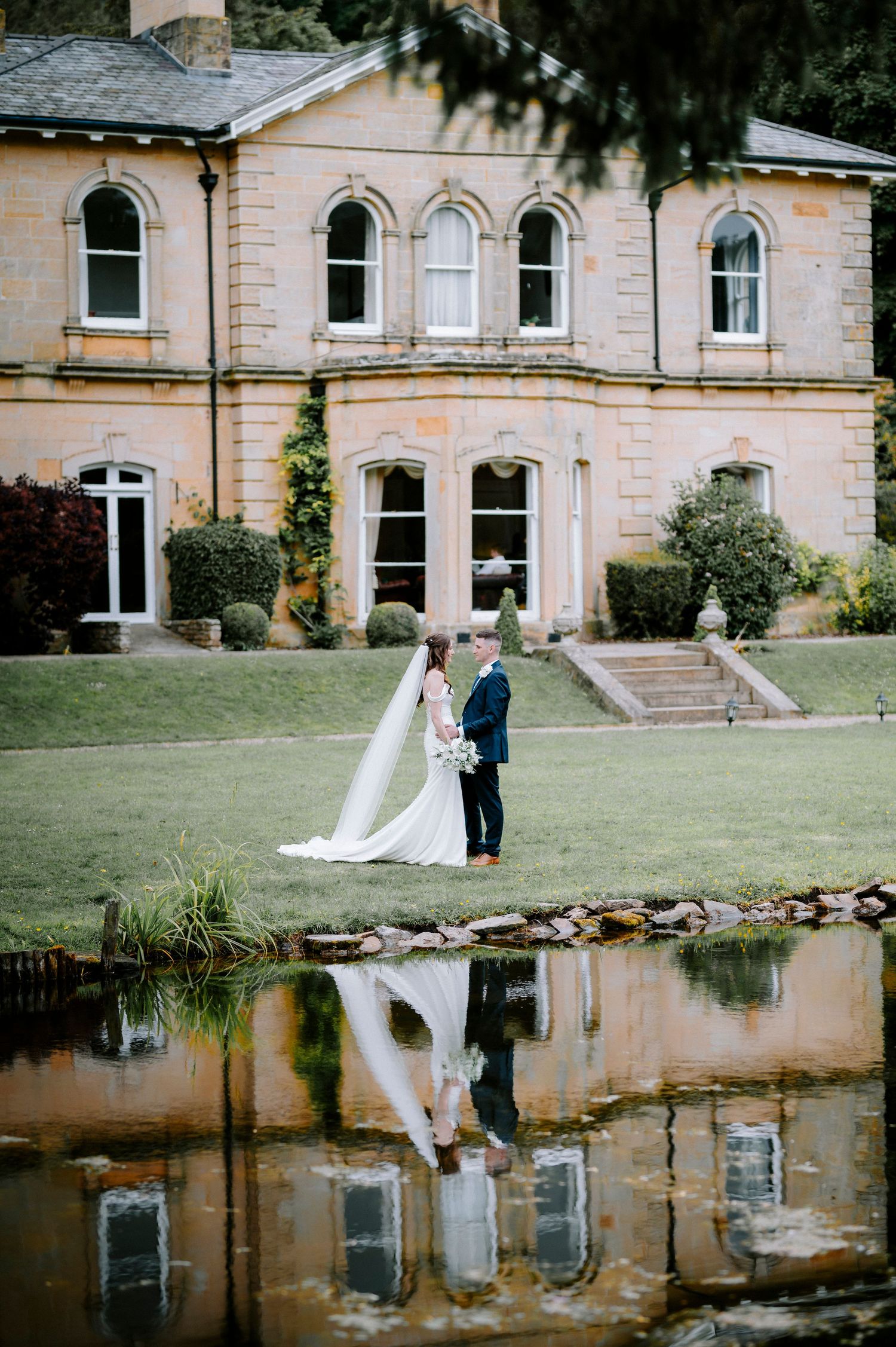 Wedding couple poses for photos in front of a grand stone mansion with reflecting pond.