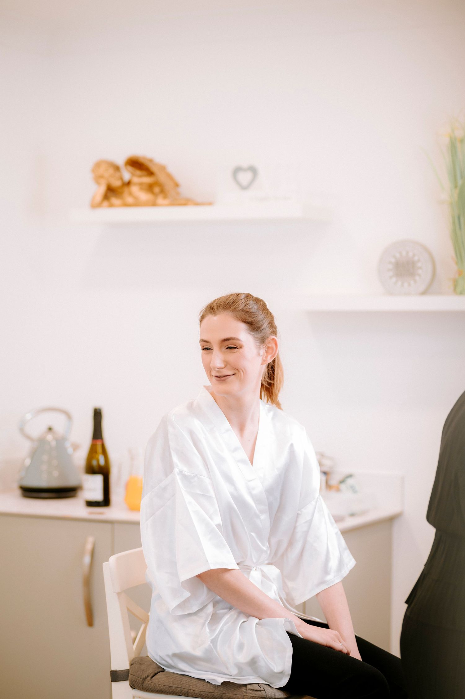 A person in a white silk robe sits on a chair during wedding preparations.