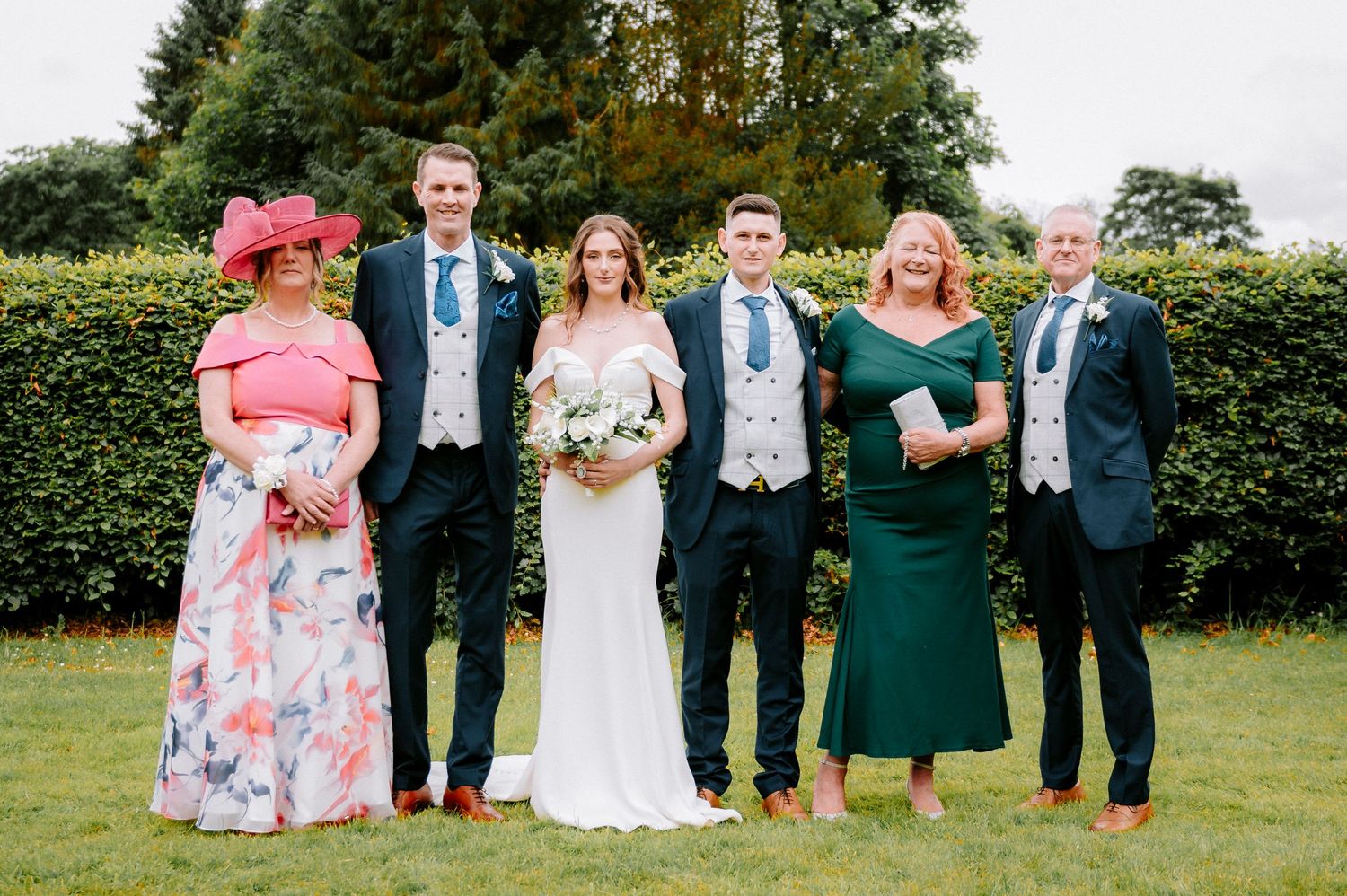 Wedding party in formal attire poses against natural outdoor backdrop.