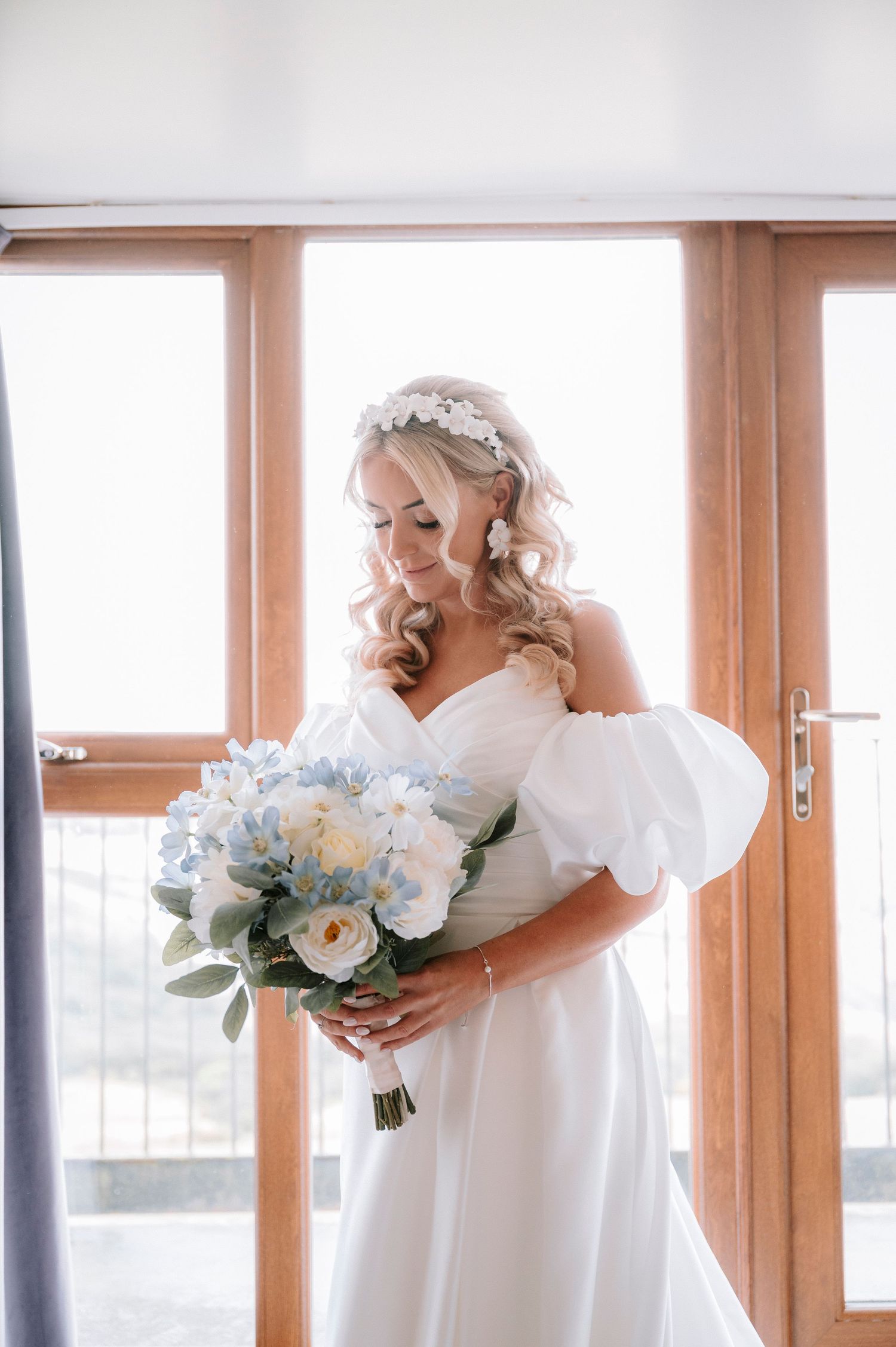 Bride in white dress with puff sleeves holds a blue and white bouquet by sunlit windows.