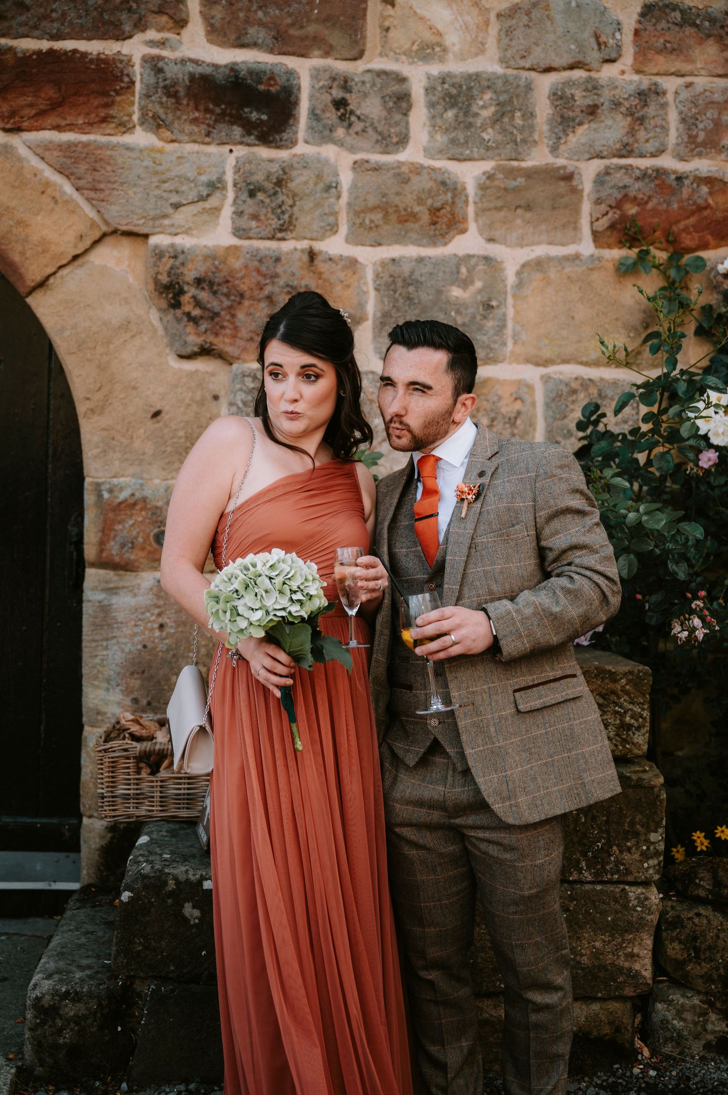Wedding party in rust-colored dresses and tweed suits posing against a stone wall.