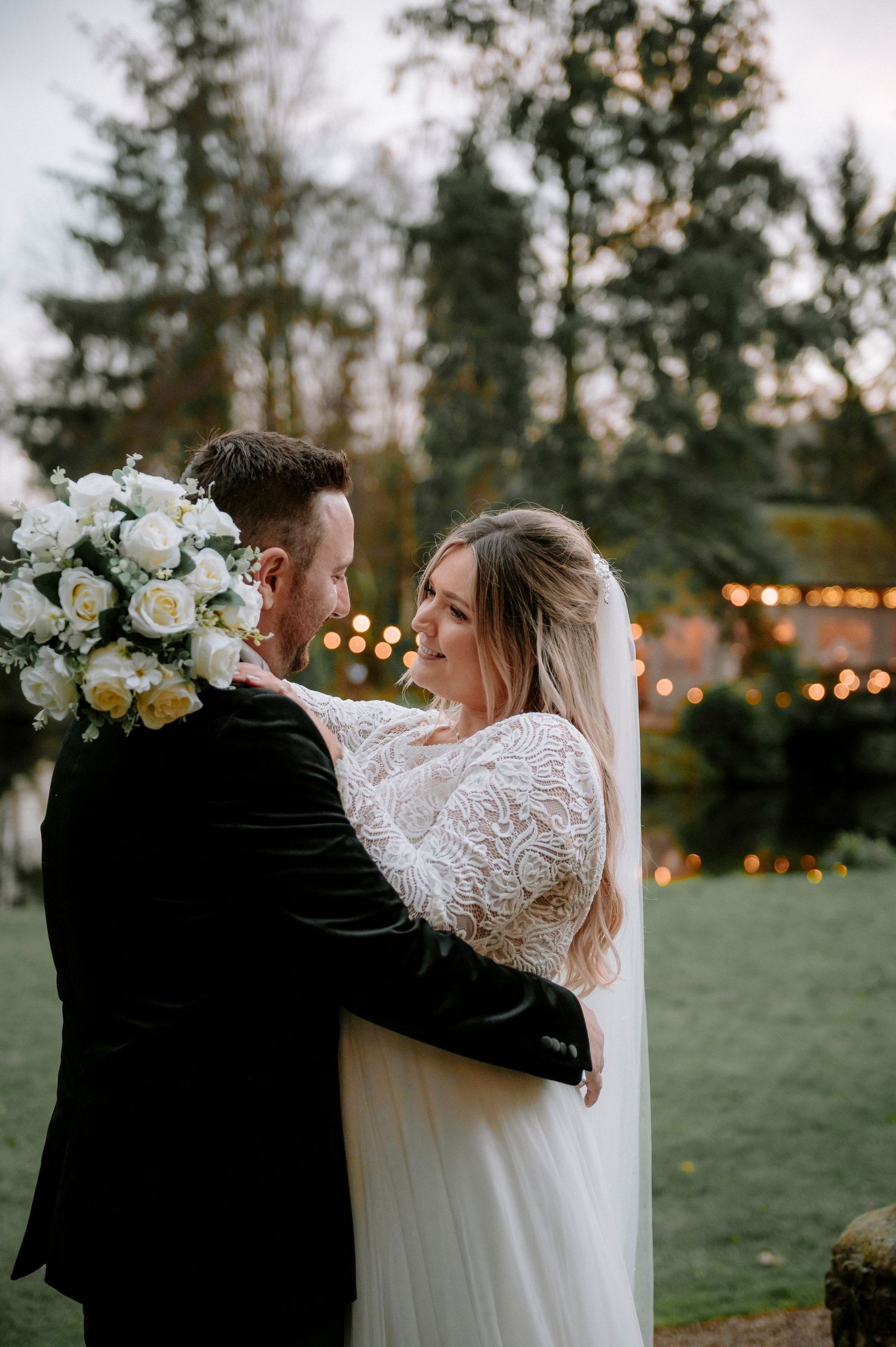 A romantic wedding couple embracing outdoors with string lights and floral decorations.
