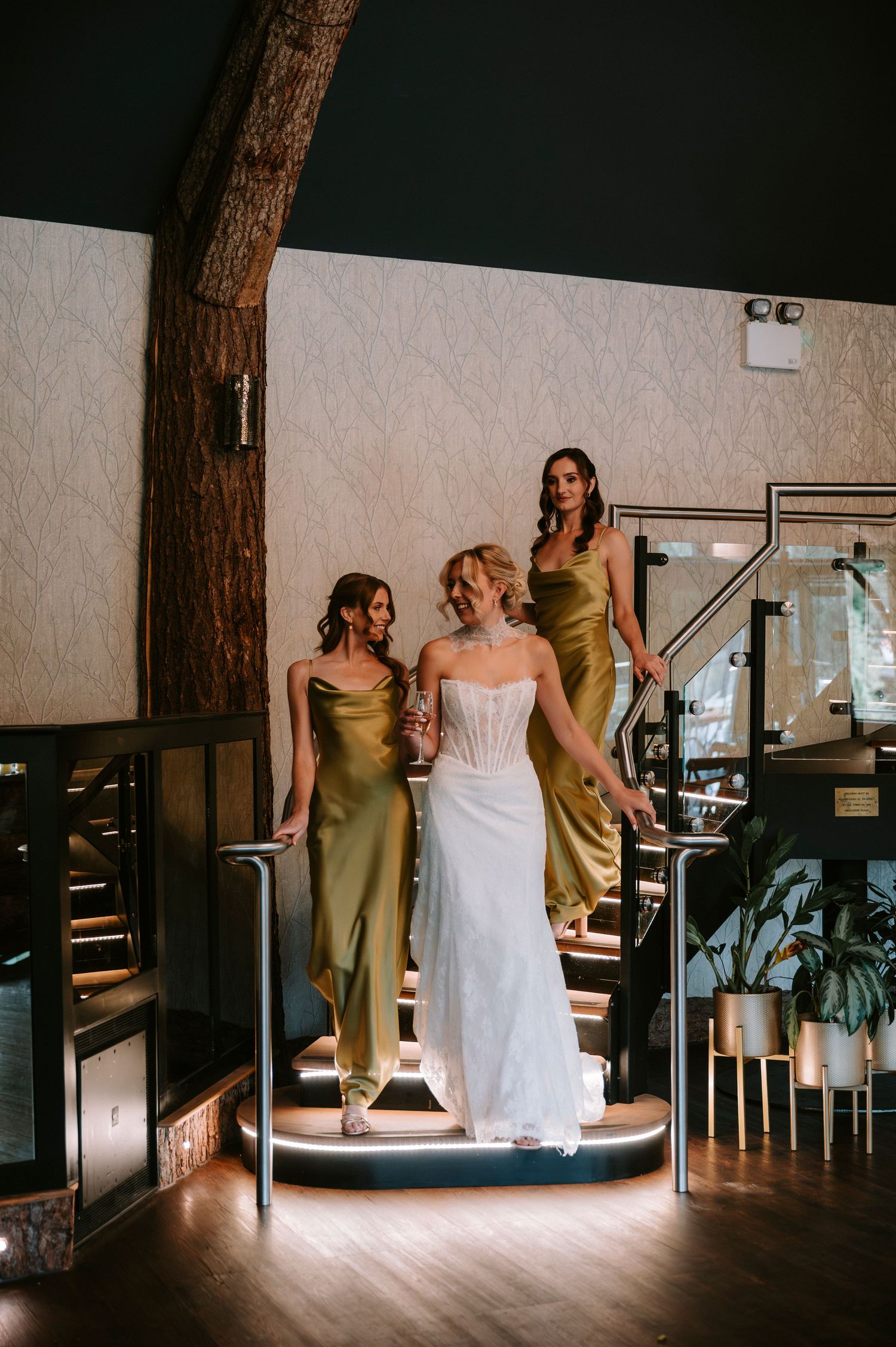Wedding party in olive green dresses and white gown gather in a dimly lit venue with rustic wooden elements.