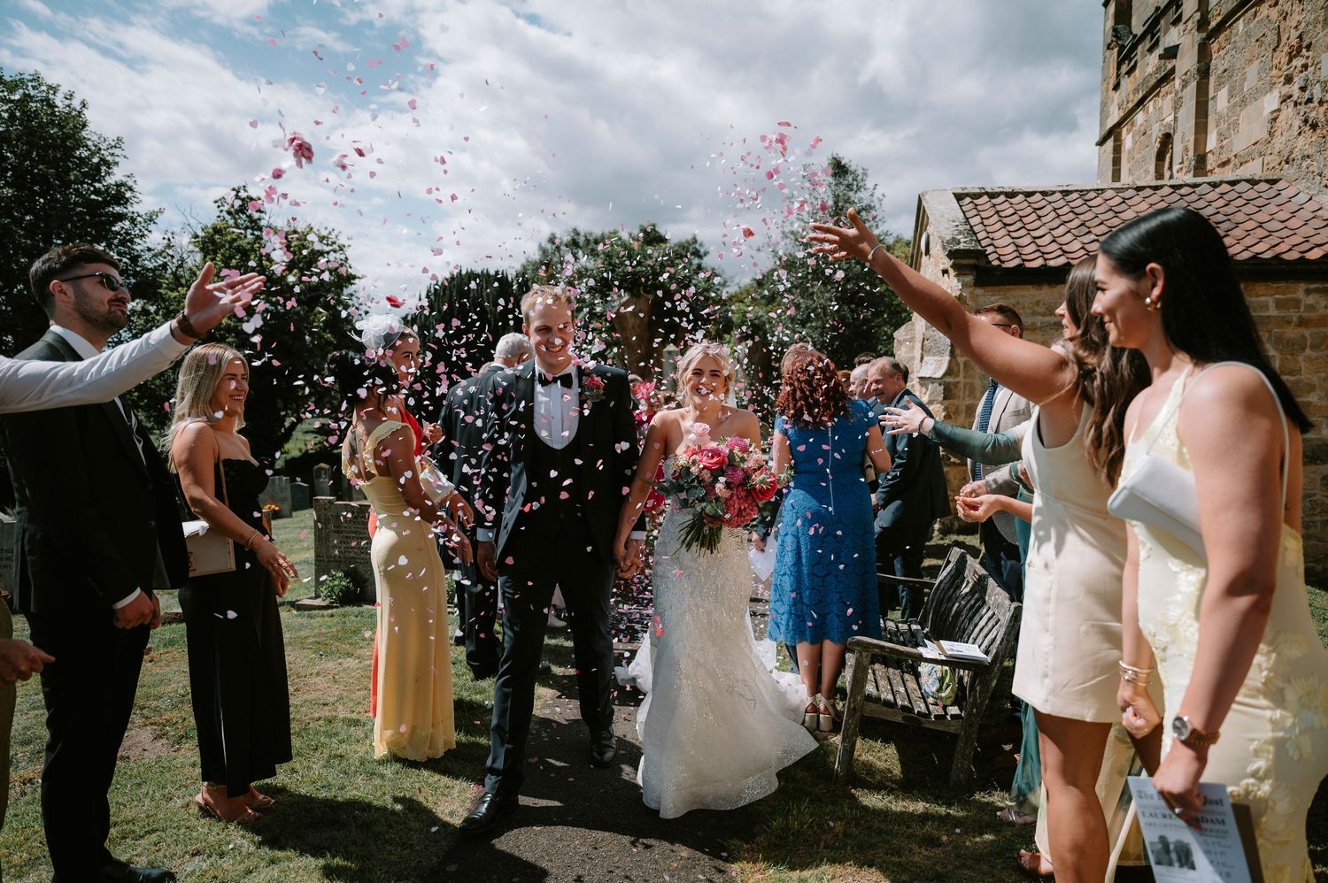 Wedding party celebrates with confetti against cloudy sky backdrop.