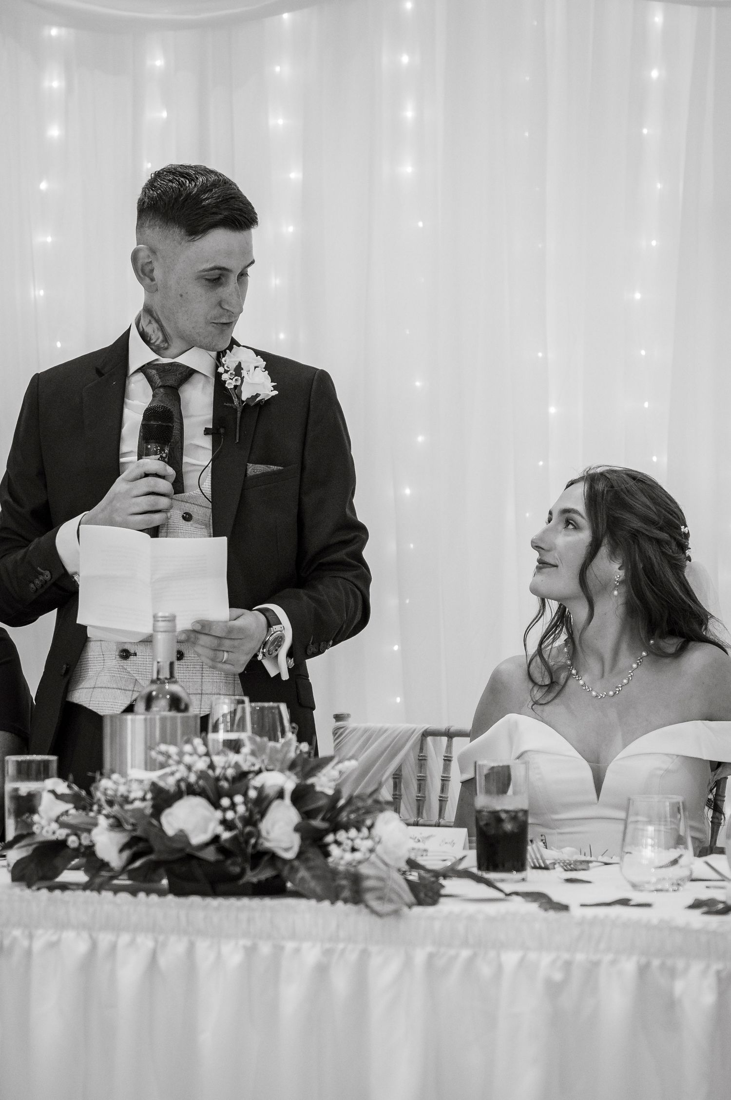Wedding couple and guests seated at head table with twinkle light backdrop during reception speeches.