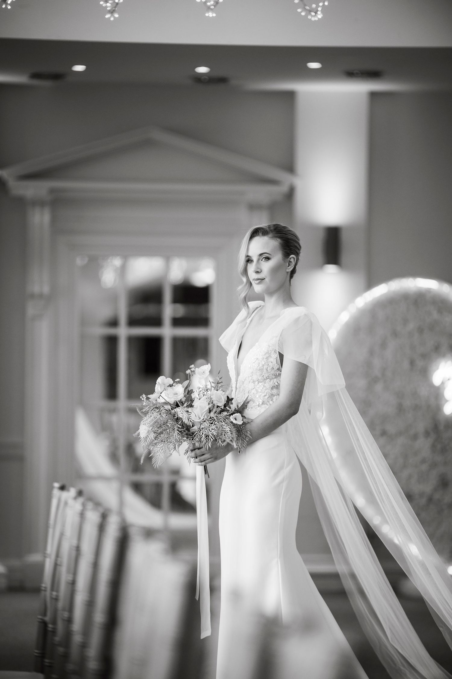 Black and white portrait of elegant bridal gown with flowing veil in classic venue setting.