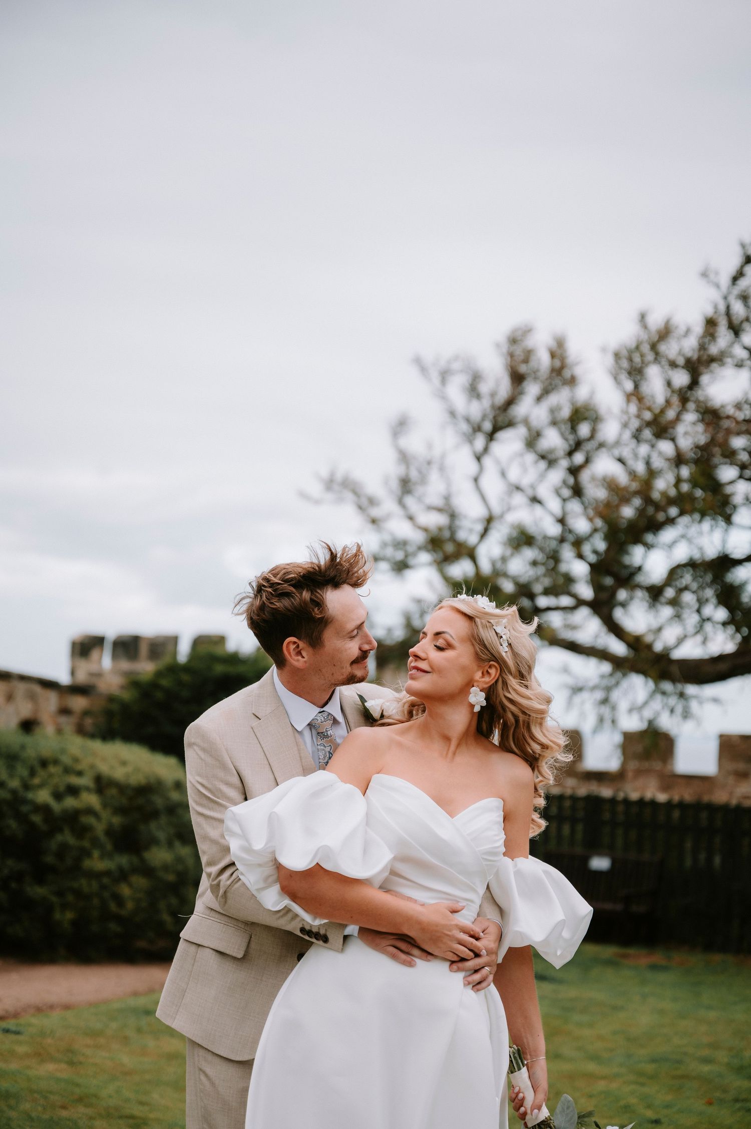 Romantic wedding couple embraces on a lawn with castle walls in the background.