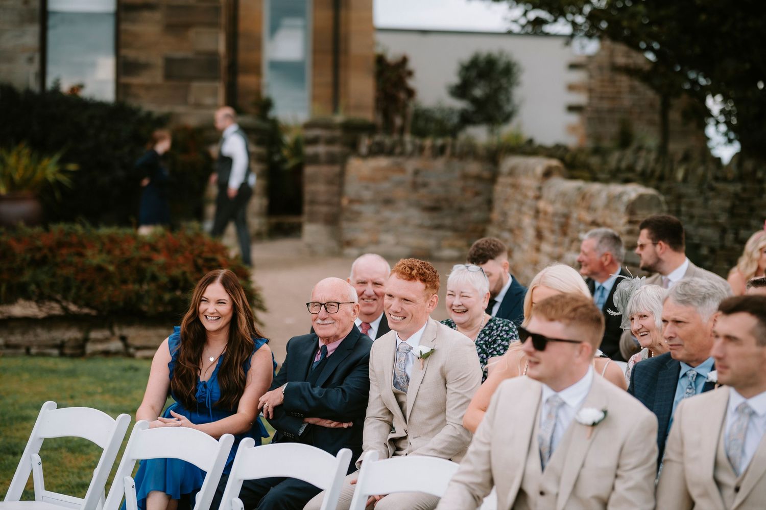 Wedding guests seated in white chairs on a lawn during an outdoor ceremony.