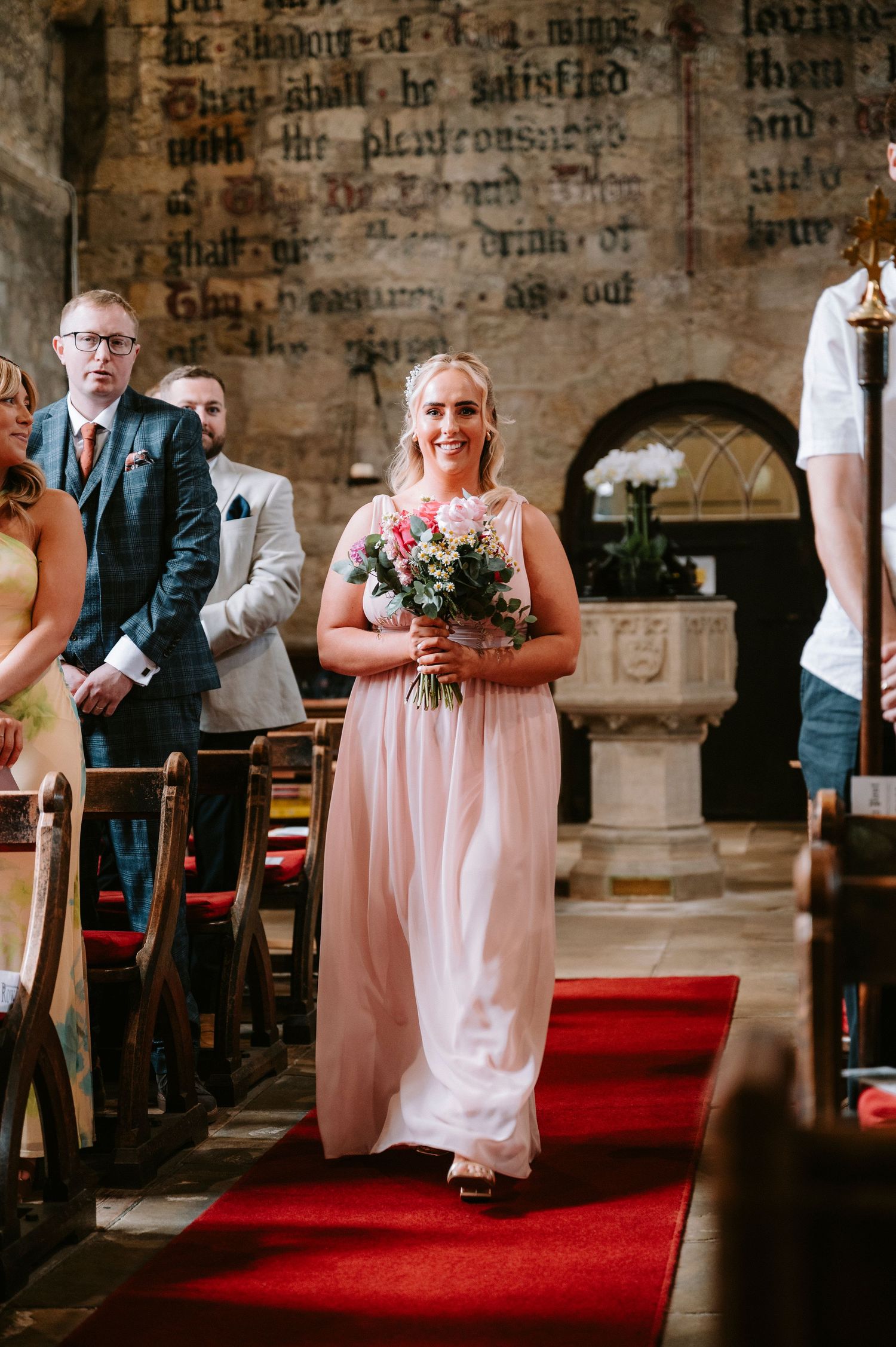 Bridesmaid in pink dress walks down red-carpeted church aisle carrying floral bouquet.