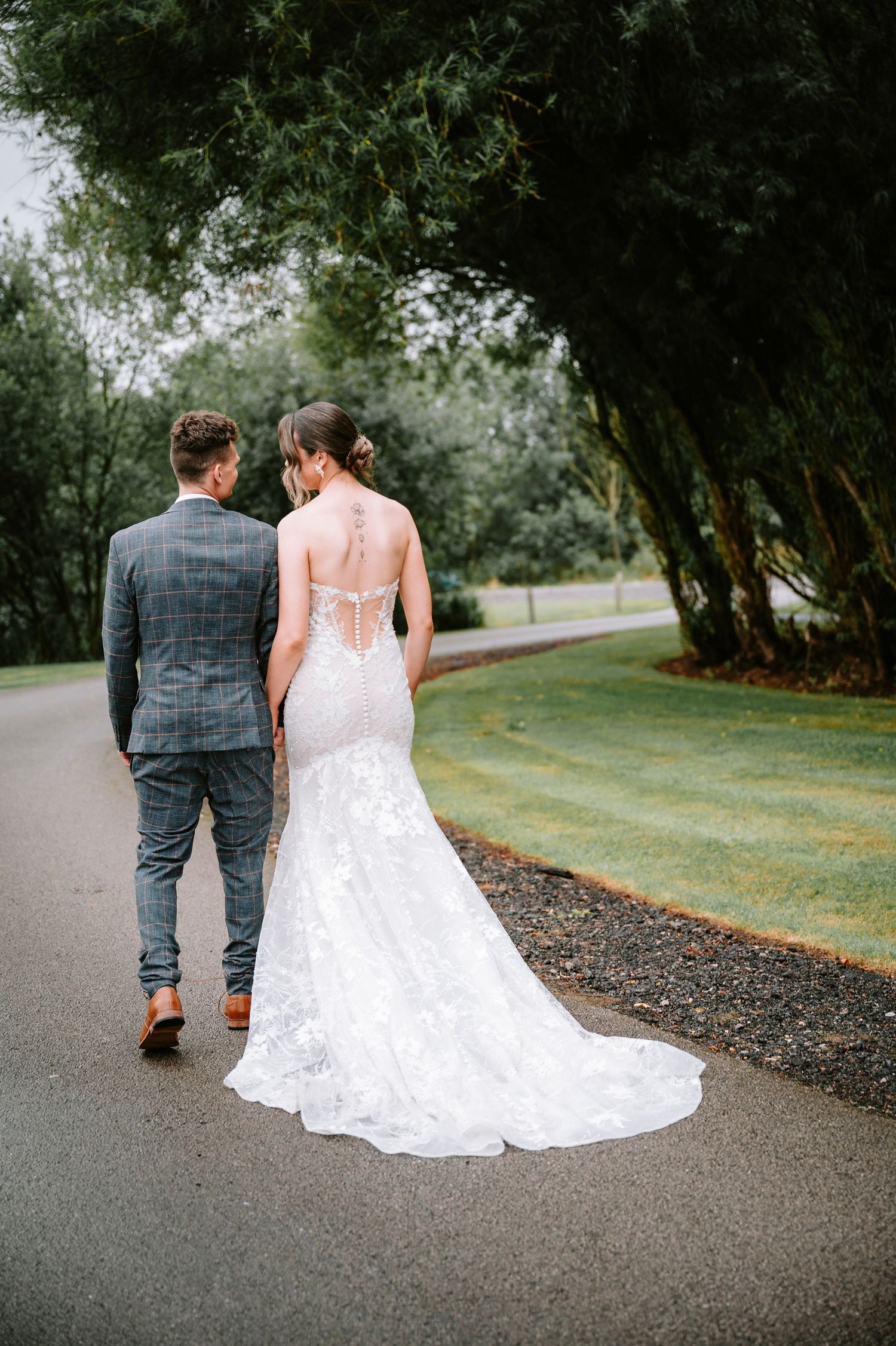 Couple walks along outdoor path with bride's lace dress trailing behind.