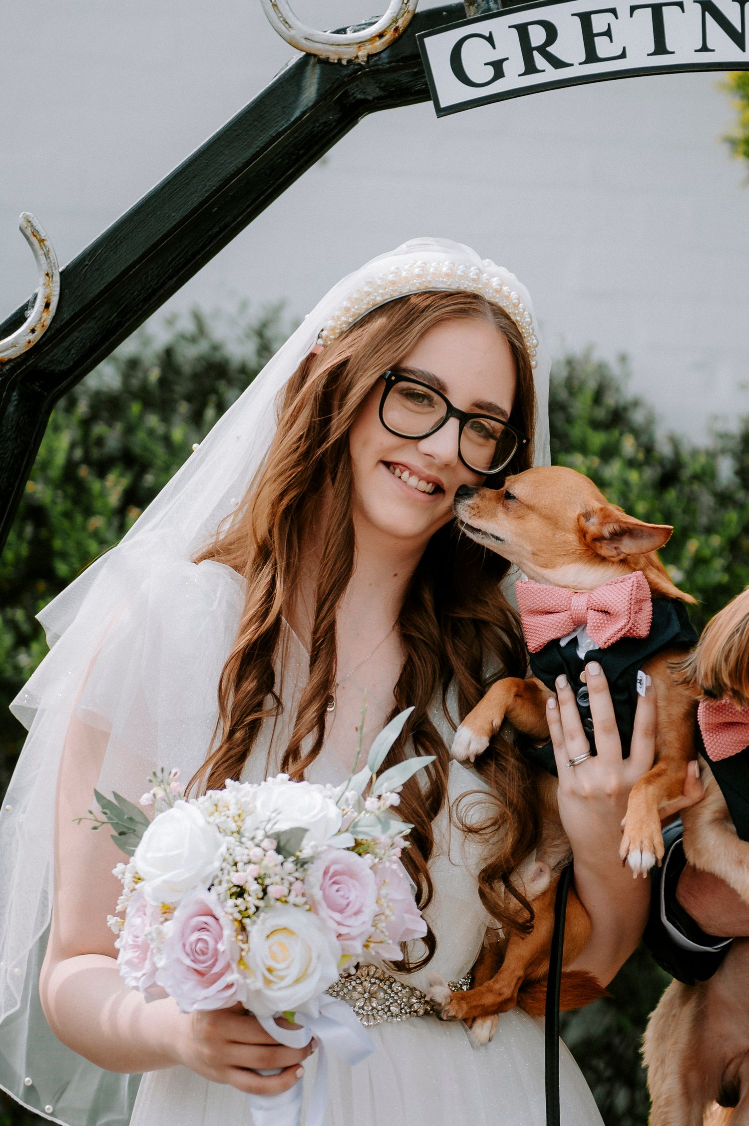 Bride in glasses holds a pink and white bouquet while sharing a moment with a dog at Gretna Green.