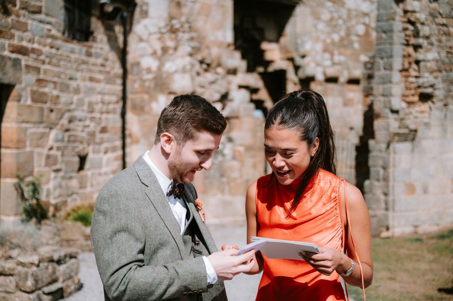 A couple in formal attire shares a moment while looking at something together near a brick wall.