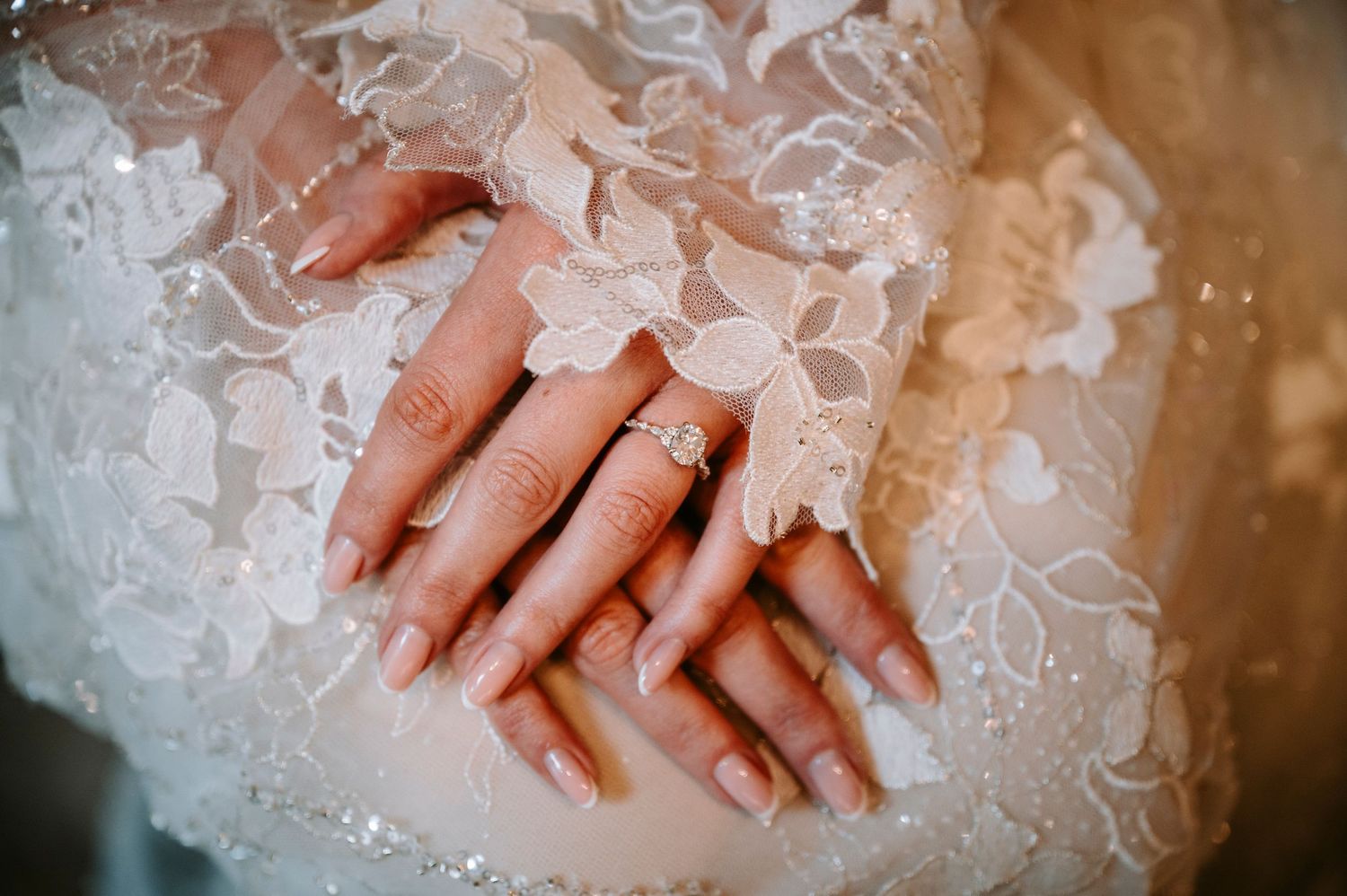 Close-up of delicate lace wedding dress details with hands showing engagement ring.