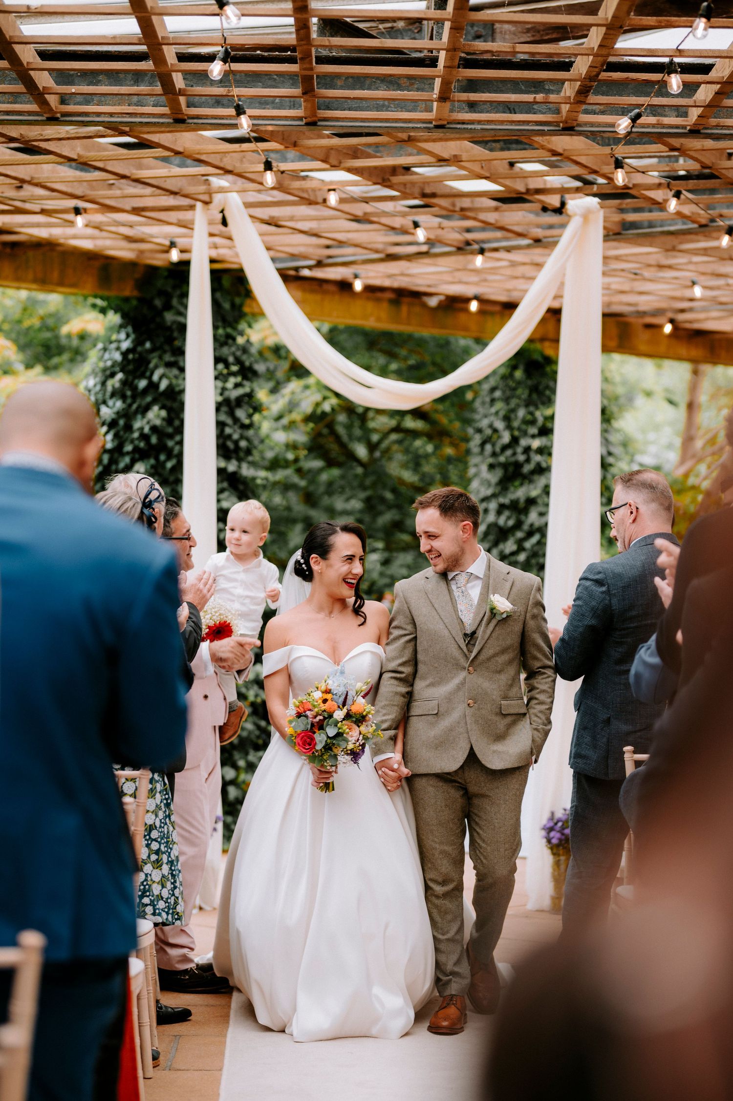 Wedding party celebrates under rustic wooden pergola with white fabric draping.