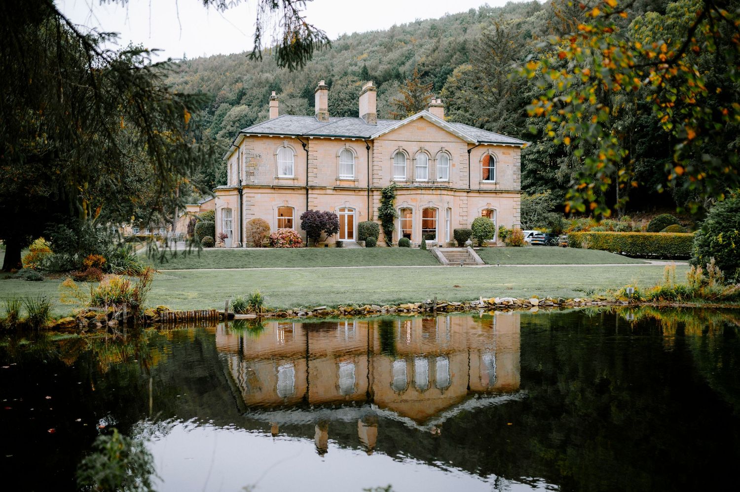 Historic mansion with peach-colored walls reflecting in a serene lake surrounded by autumn trees.