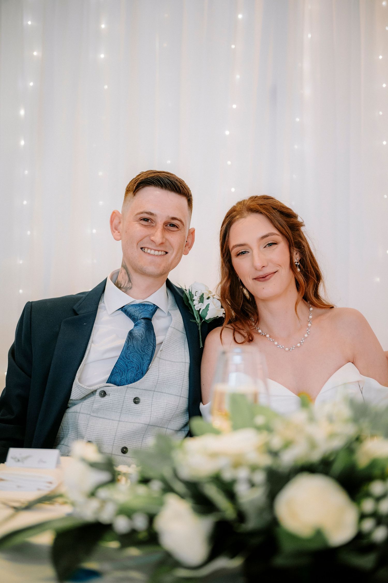 Newlyweds seated at reception table with white floral arrangements and twinkling backdrop.