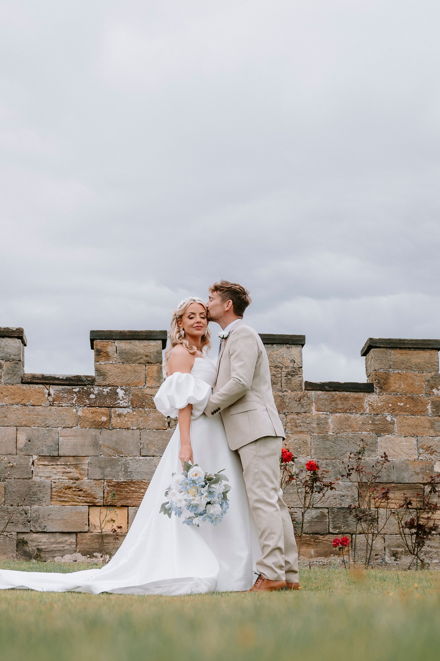 Newlyweds embrace against stone castle wall with cloudy sky and red flowers in background.