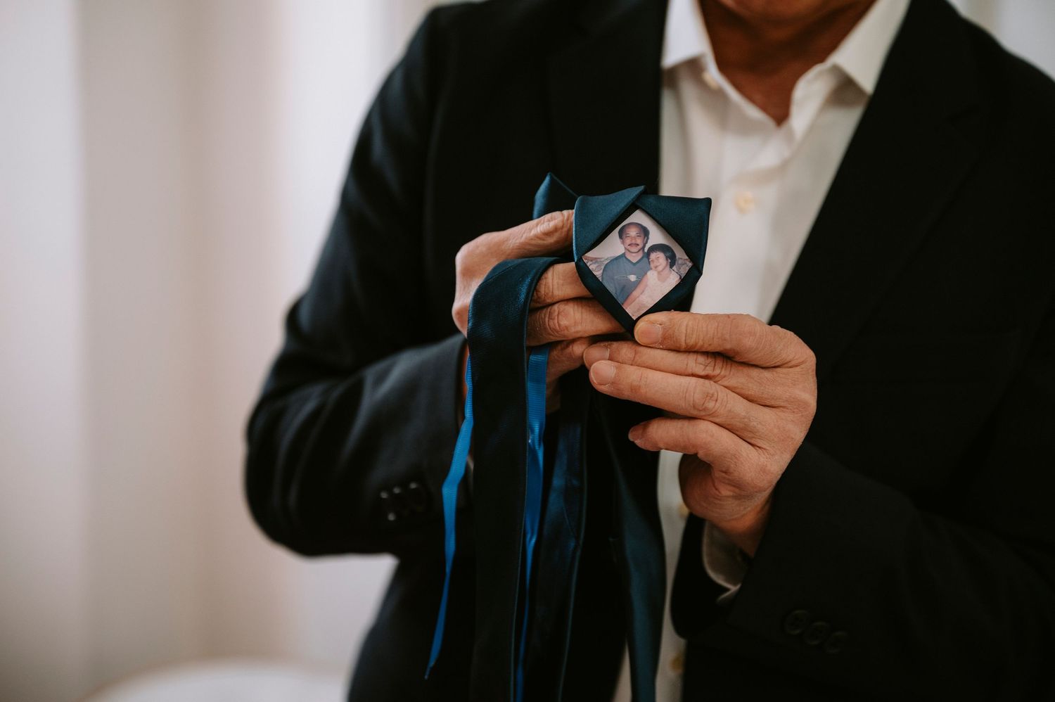 Person in black suit holding playing cards with blue ribbon against light background.