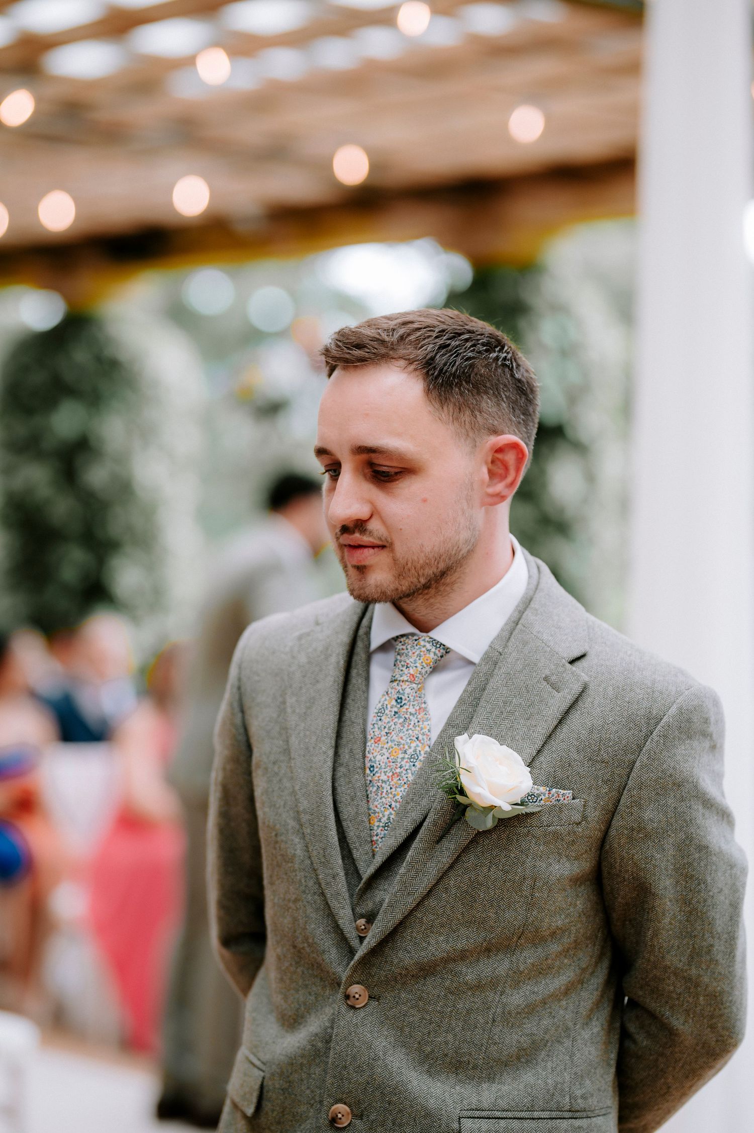 Groom in a gray suit with floral tie smiles during outdoor wedding ceremony.