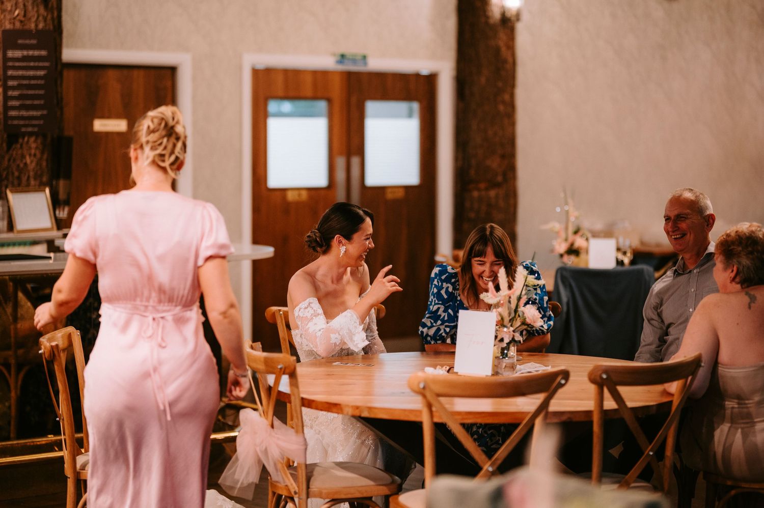 People in formal attire mingle and dance at a wedding reception in a rustic venue with wooden decor.