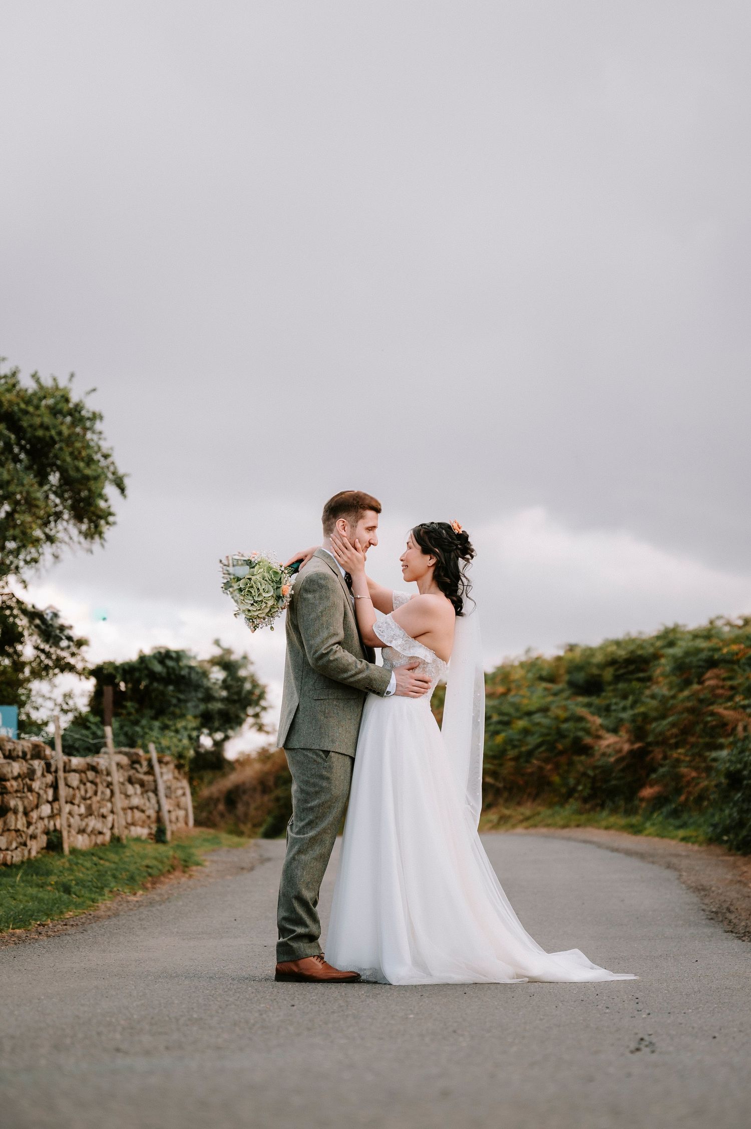 Newlyweds share romantic moments on a country road with stone walls and greenery in the background.